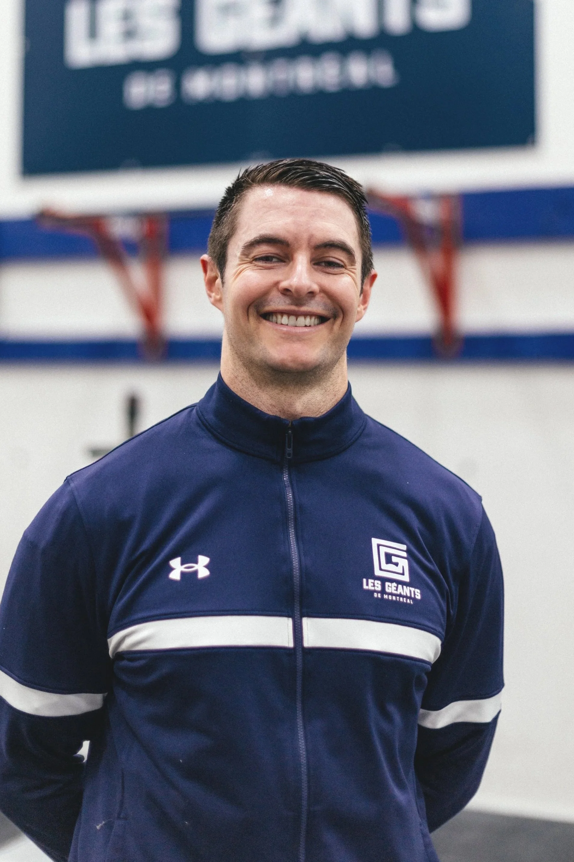Un homme souriant portant un uniforme de sport bleu avec un logo 'Les Géants de Montreal' et le logo Under Armour, dans un gymnase avec un panneau flou en arrière-plan.
