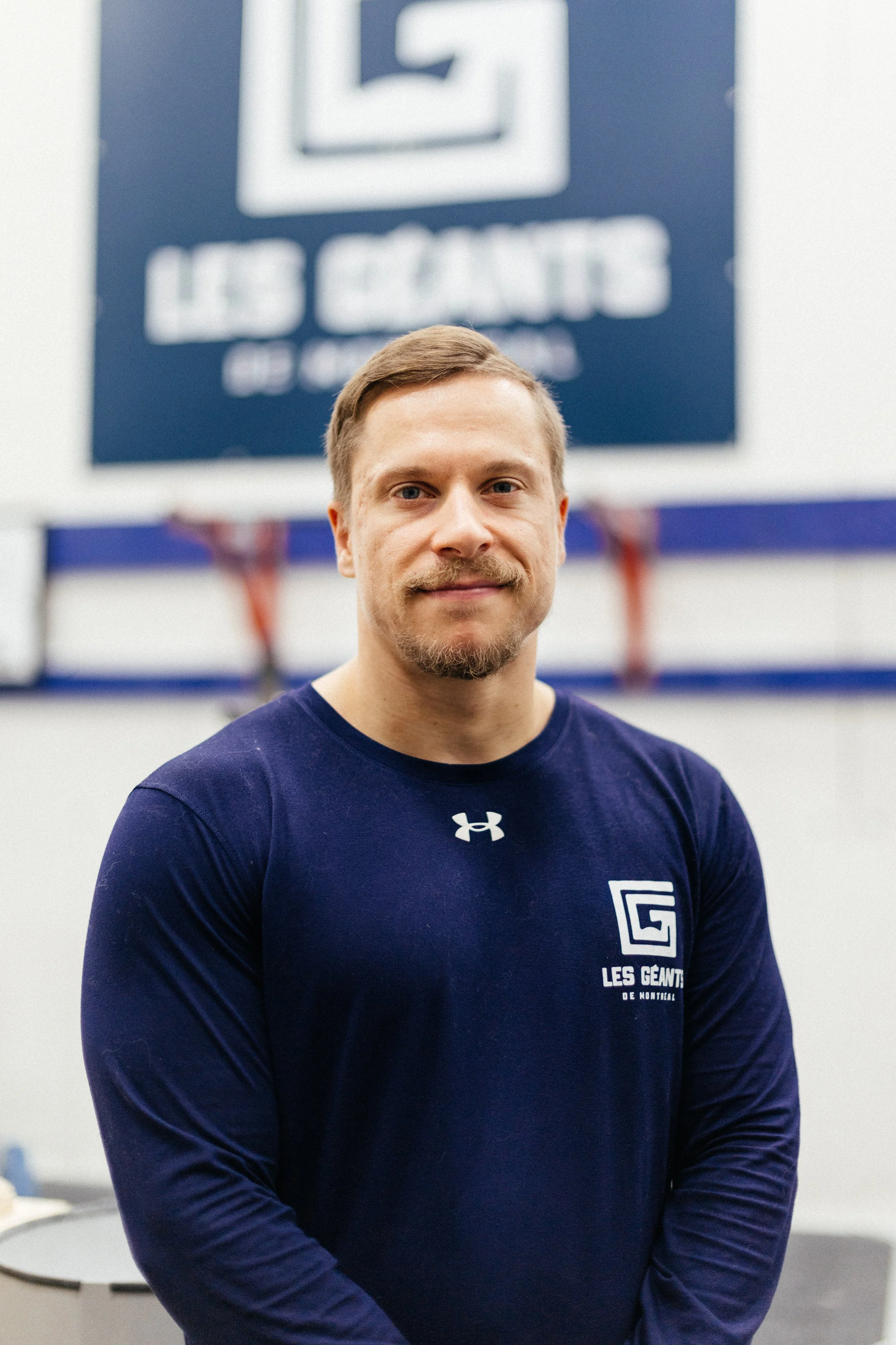 Un homme portant un t-shirt bleu foncé avec un logo et le texte 'Les Géants de Montréal' se tient devant un panneau indiquant un espace réservé pour une pharmacie dans une salle d'entraînement ou de sport.
