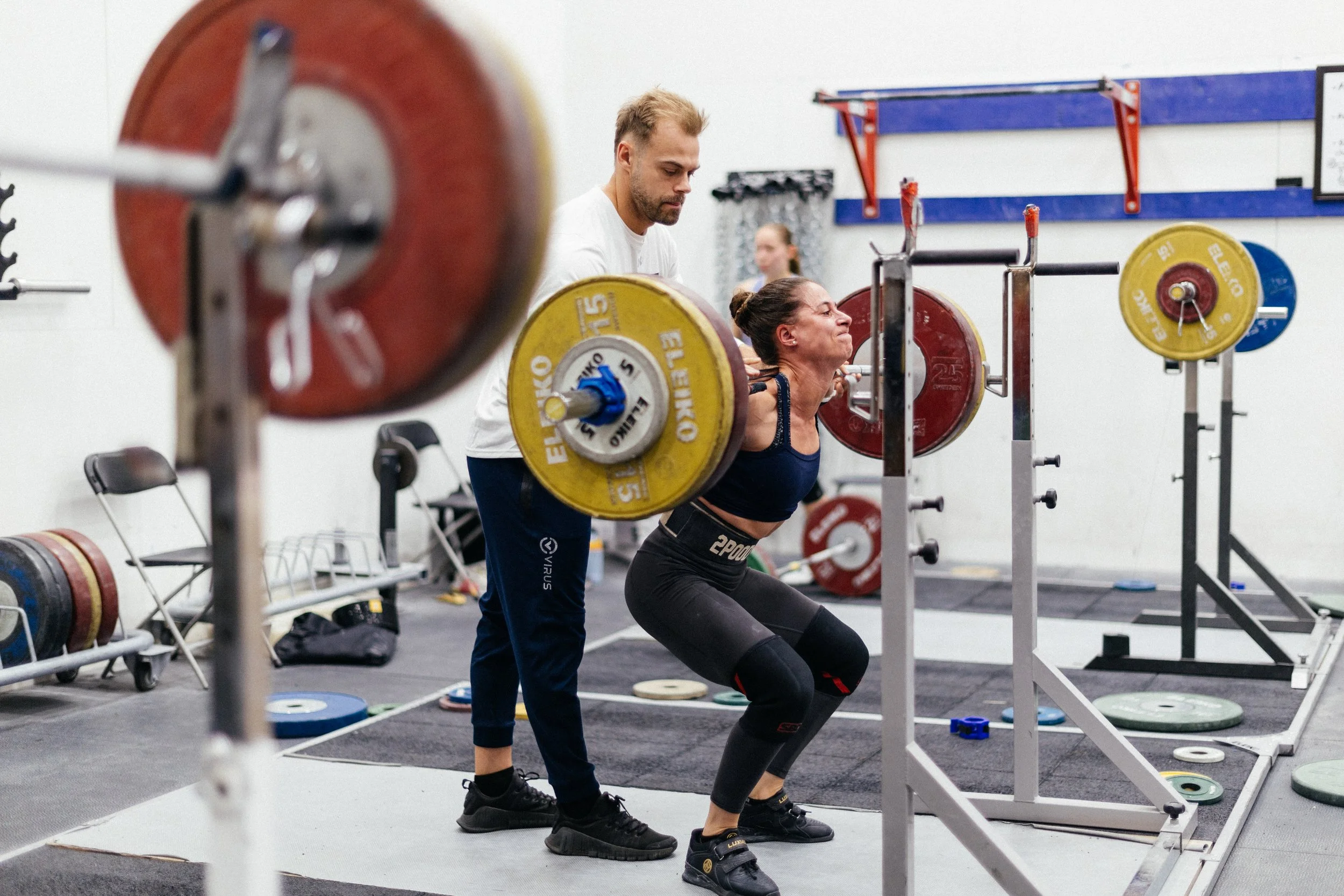 Une femme soulève une barre de poids sous la supervision d'un entraîneur dans une salle de sport.