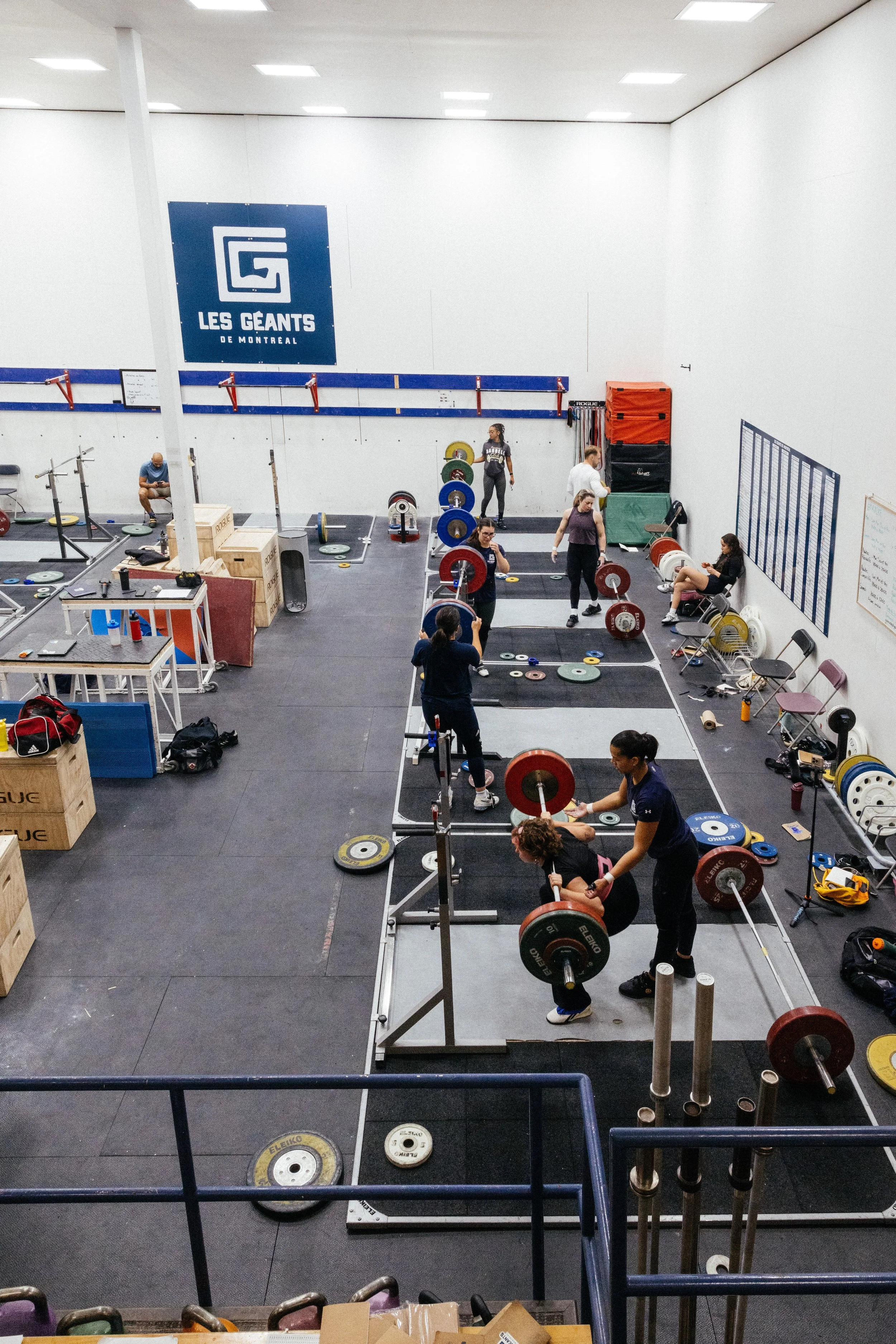 Une salle d'entraînement de gymnastique ou haltérophilie avec plusieurs personnes lèveurs de poids, équipements de musculation, et un grand panneau avec le logo 'Les Géants de Montréal'.