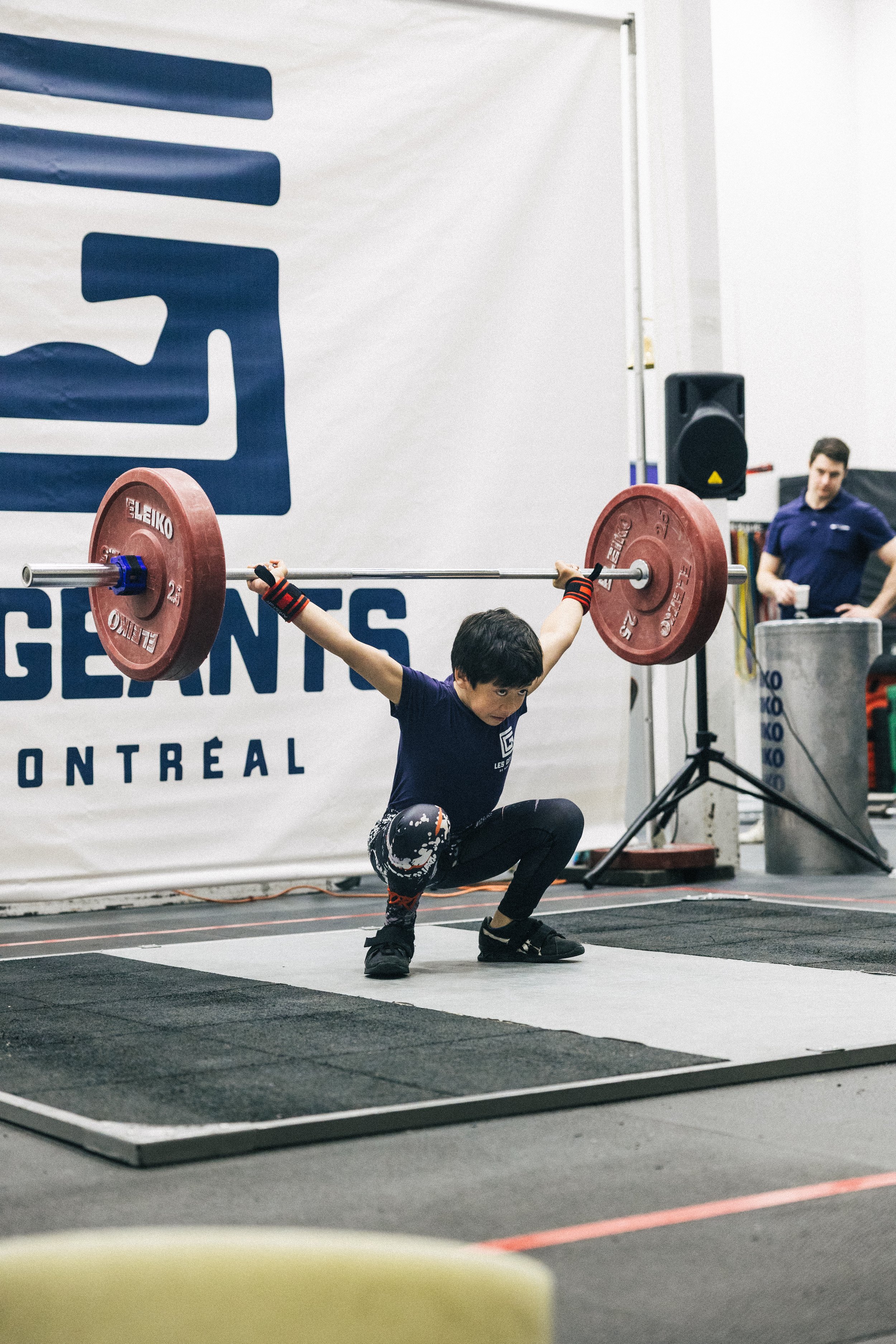 Un jeune athlète effectue un squat avec une barre chargée lors d'une compétition, avec un spectateur en arrière-plan dans une salle intérieure.