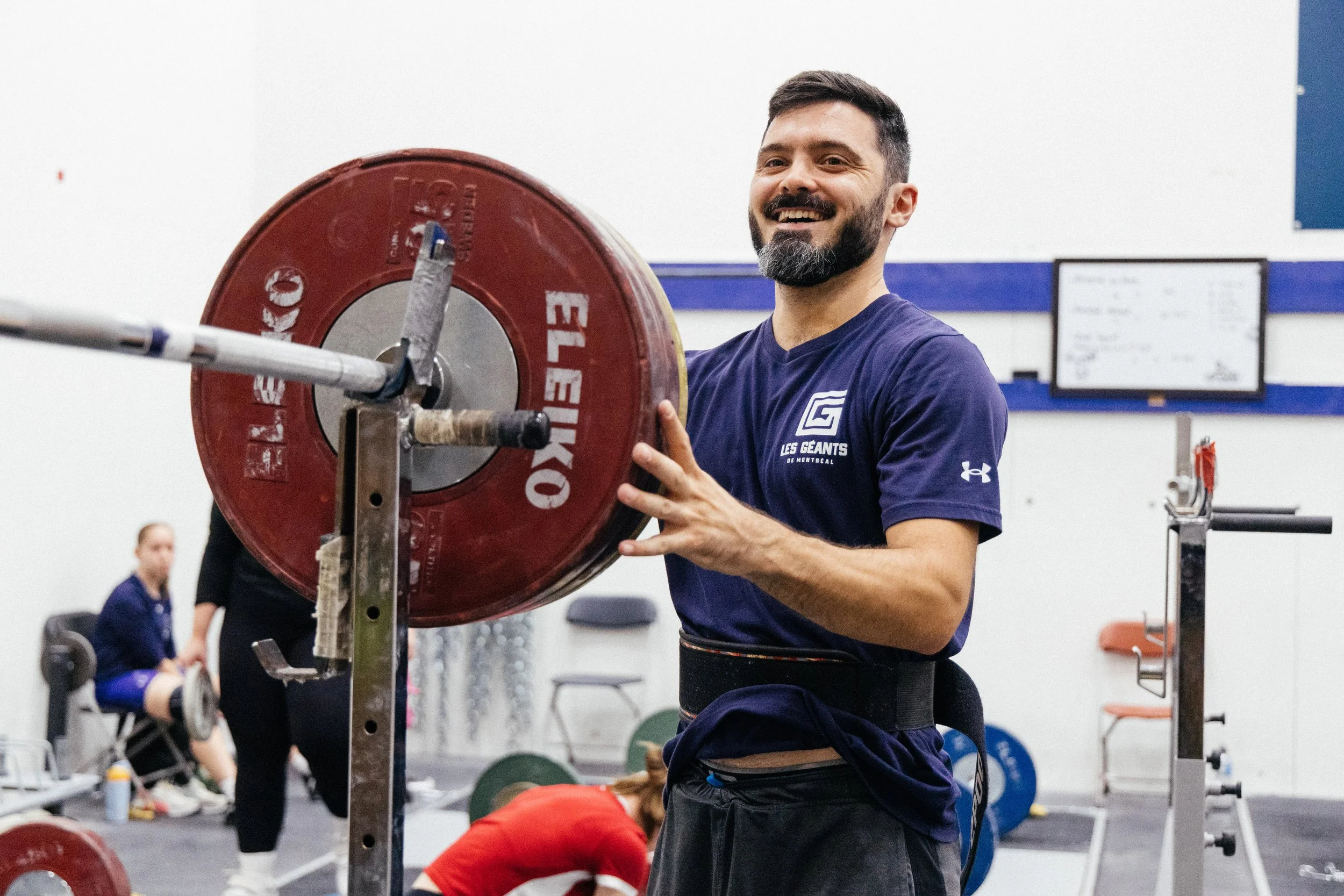 Un homme souriant en t-shirt bleu, en salle de musculation, soulève des poids sur un rack de musculation.
