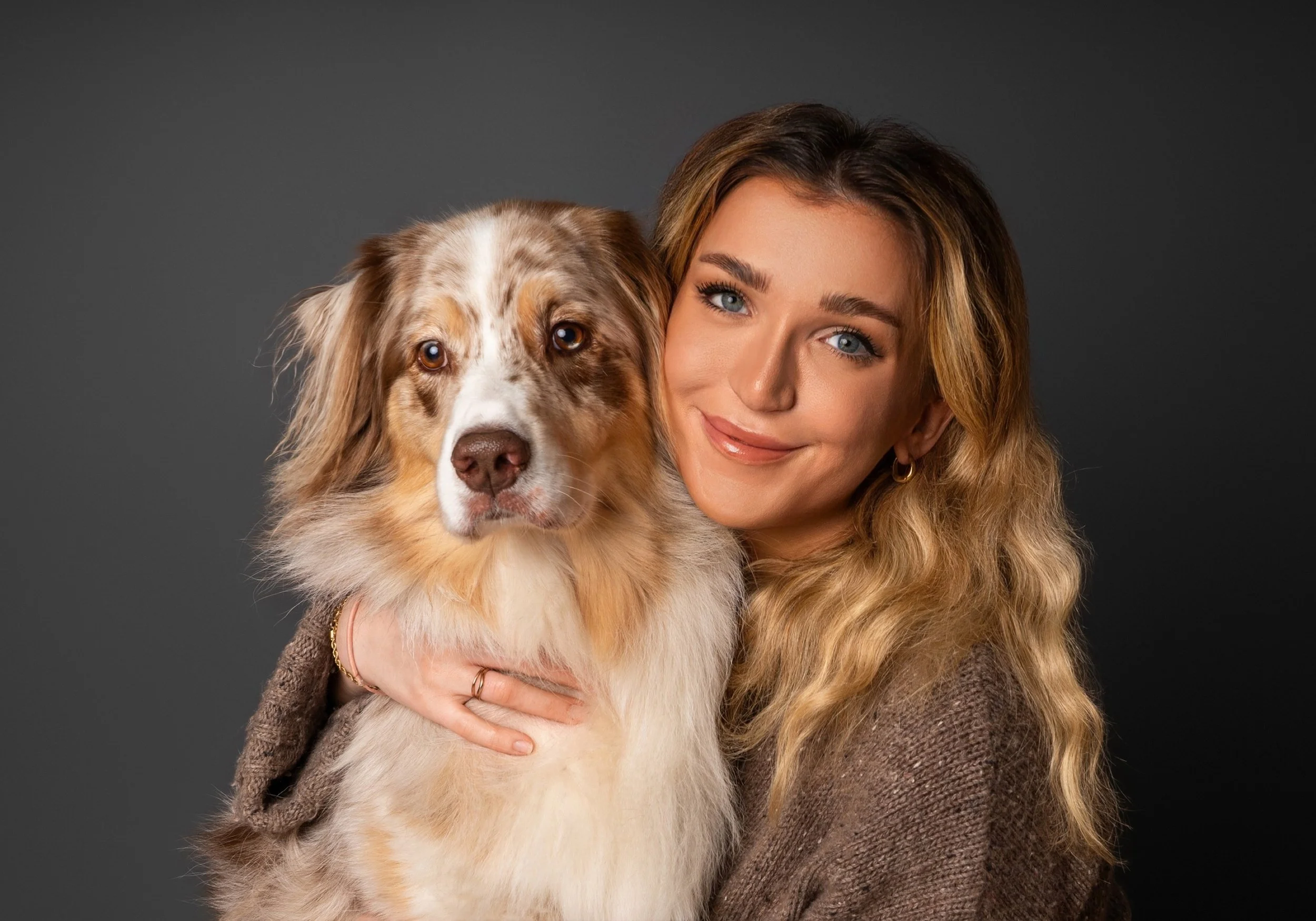 A woman with long wavy blond hair and blue eyes smiling while holding an Australian Shepherd dog with a multicolored coat against a dark background.
