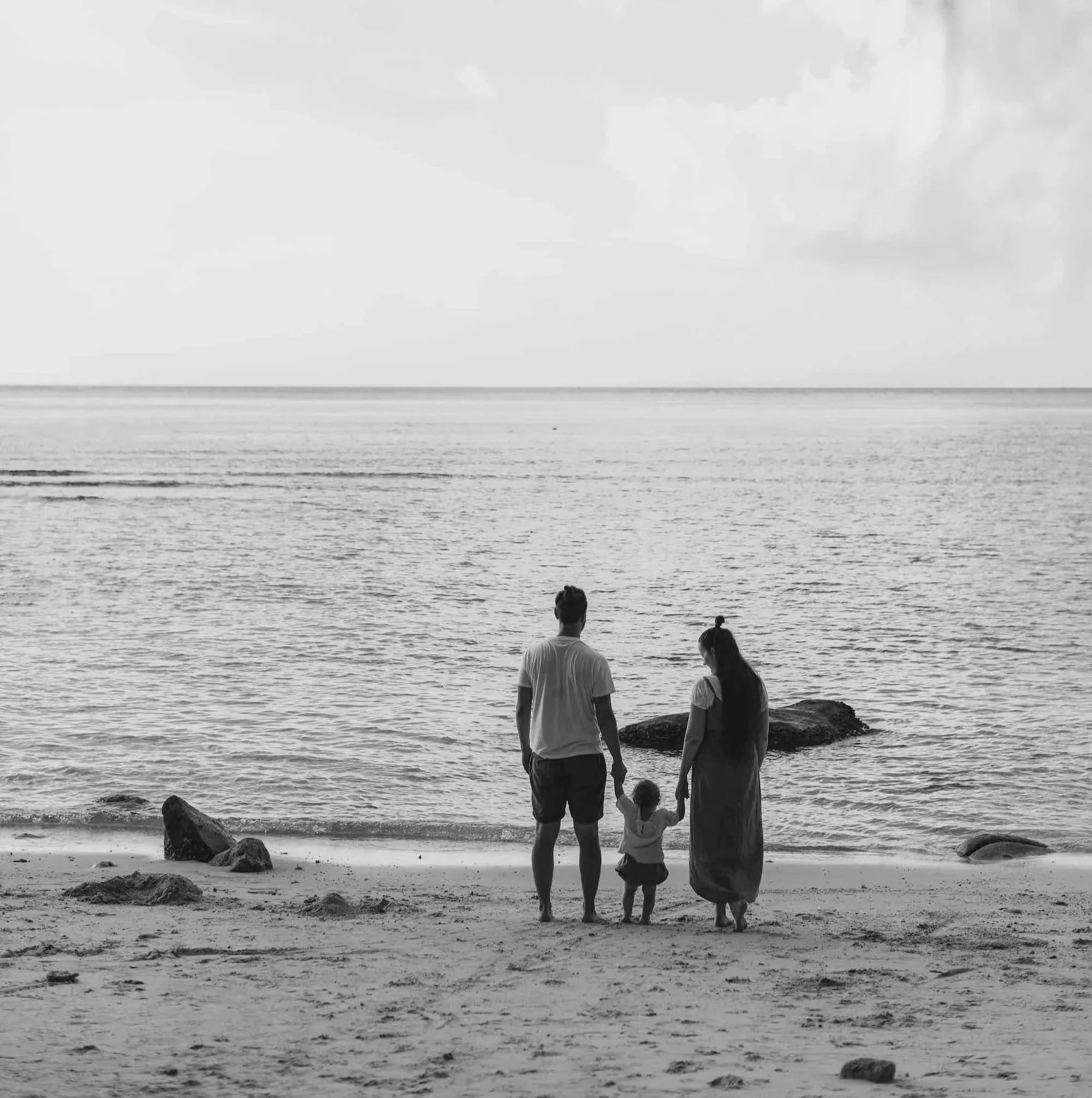 A black and white photo of a family of three holding hands and walking on a beach near the water. The man and woman are adults, and there is a small child between them. The ocean is visible in the background with a few rocks near the shore.