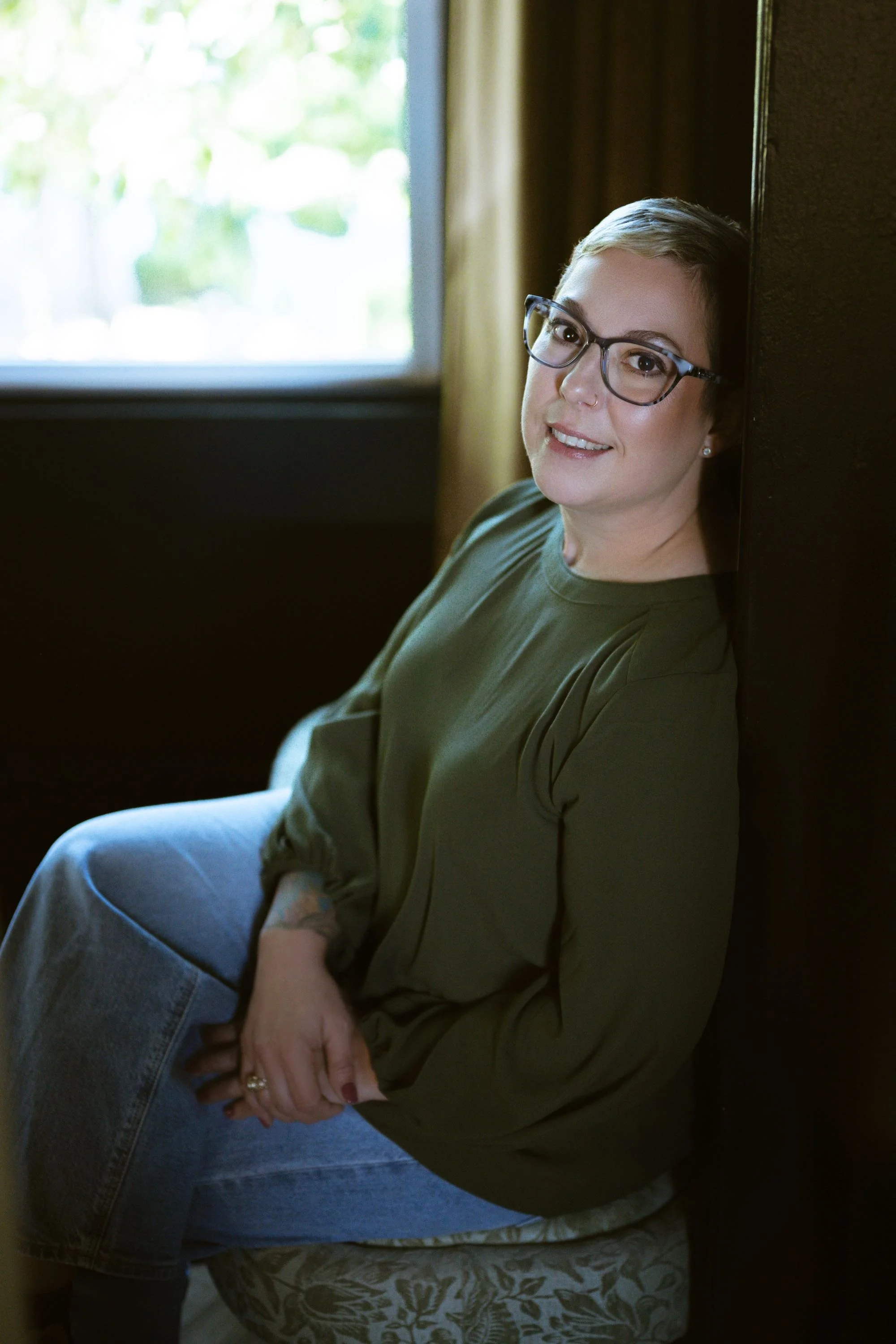 A woman with short hair, glasses, and earrings sitting on a patterned cushion, leaning against a dark wall near a window with greenery outside, wearing a green blouse and blue jeans.