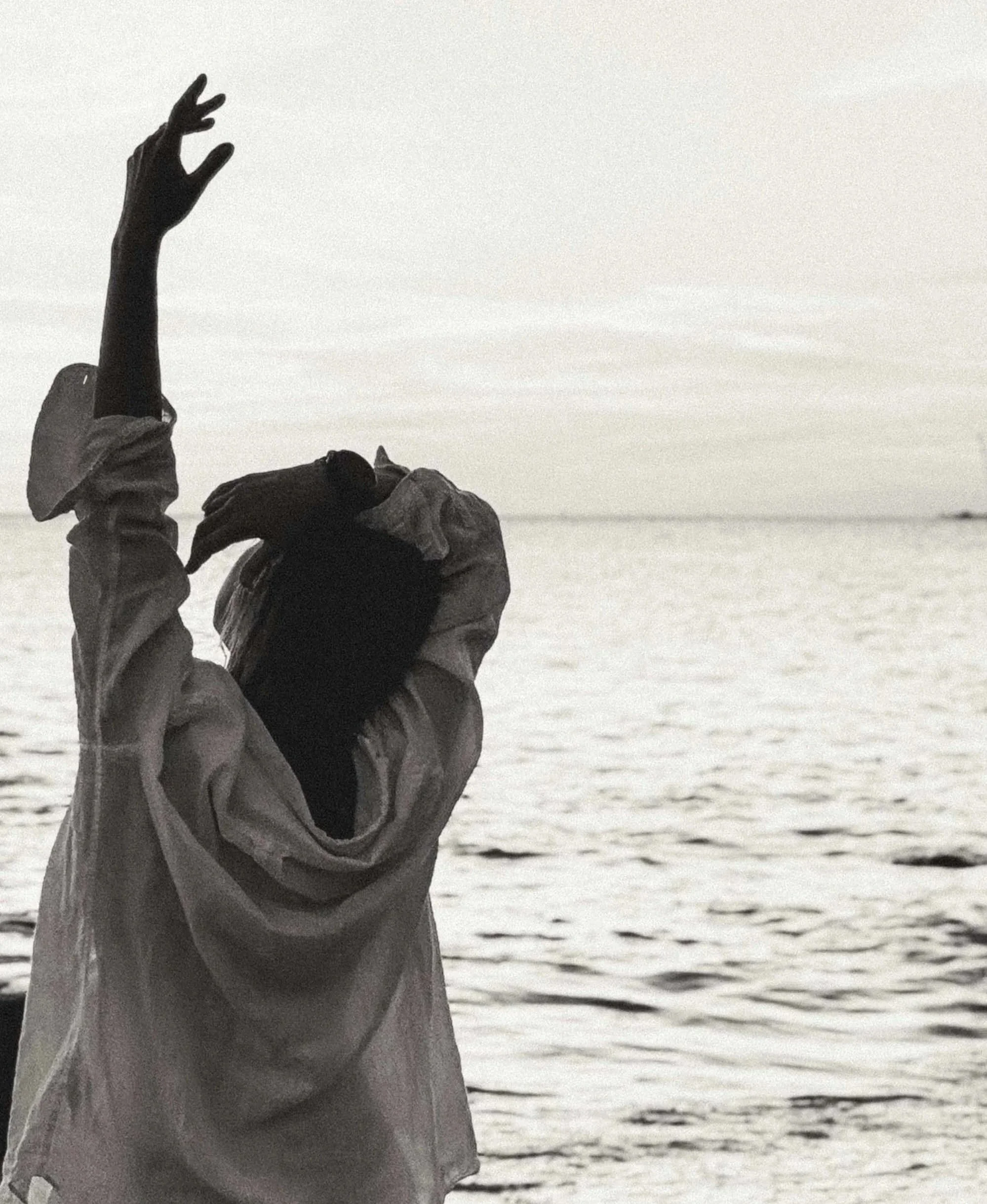 A person standing near the water at sunset, with one arm raised and head tilted back, wearing a jacket. The image is in black and white.