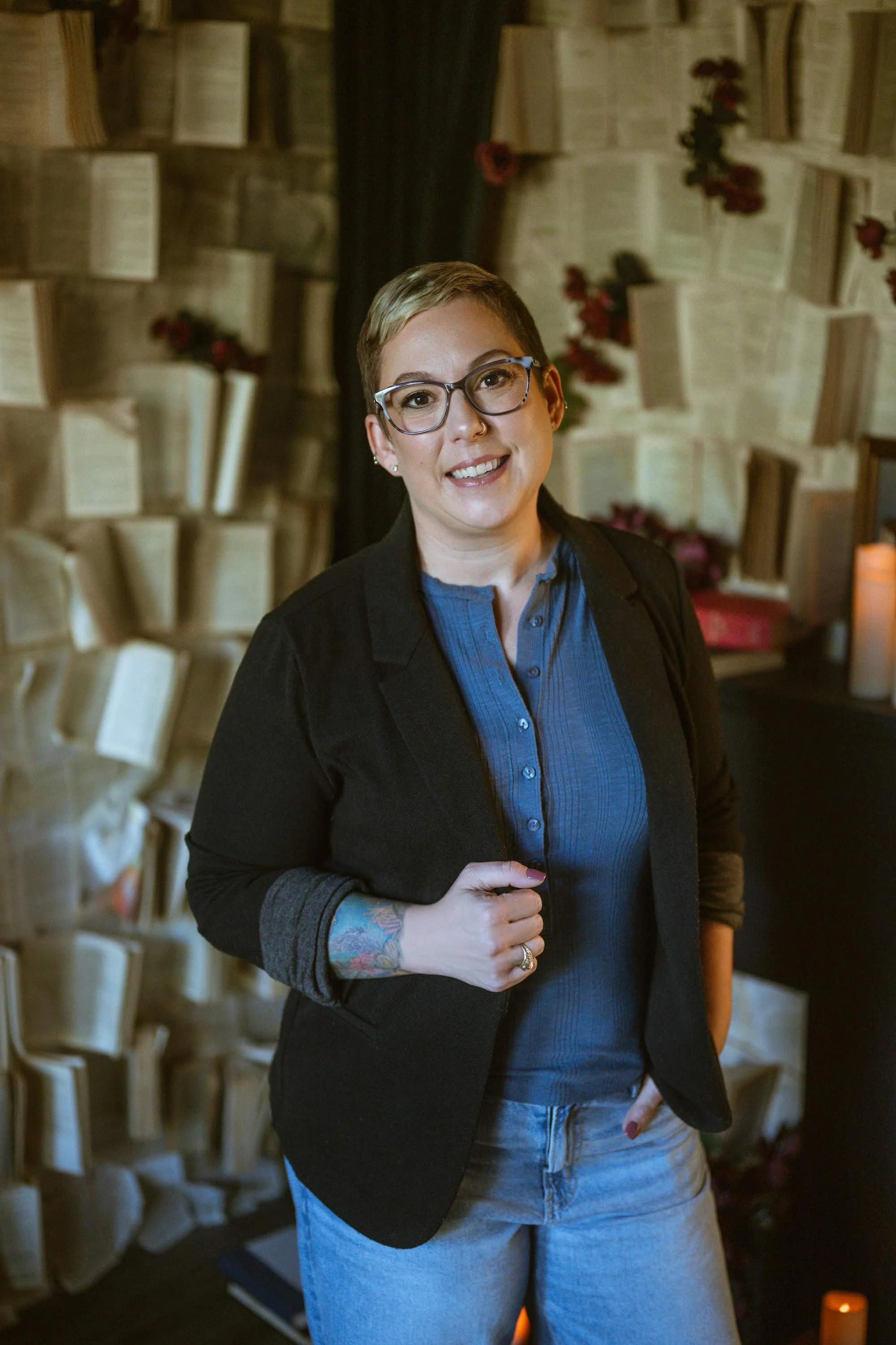 A woman with short hair, glasses, and a tattoo on her arm, wearing a black blazer and blue top, standing indoors with a background of open books and red flowers.