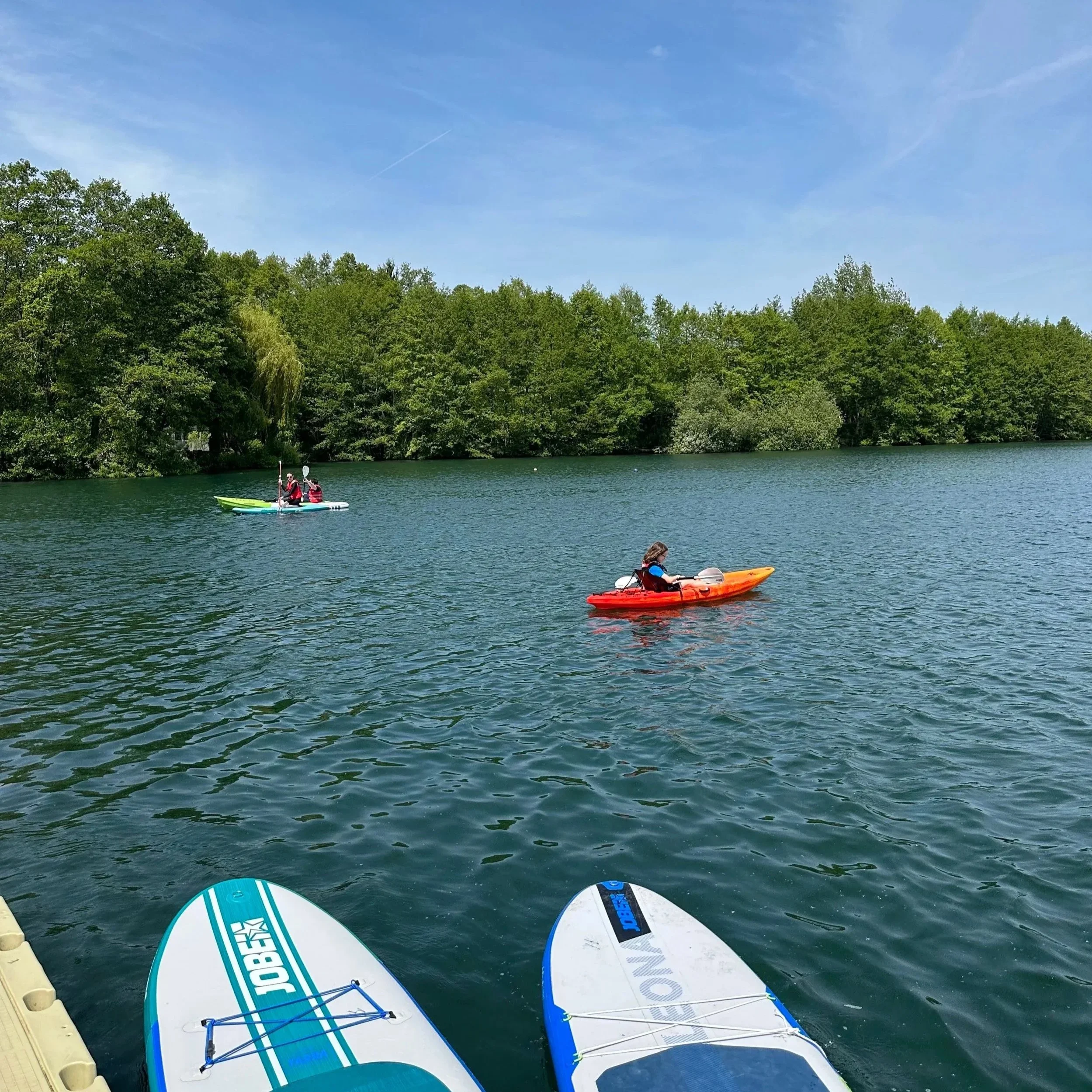 People kayaking on a body of water with lush green trees in the background, and paddleboards visible at the bottom of the image.