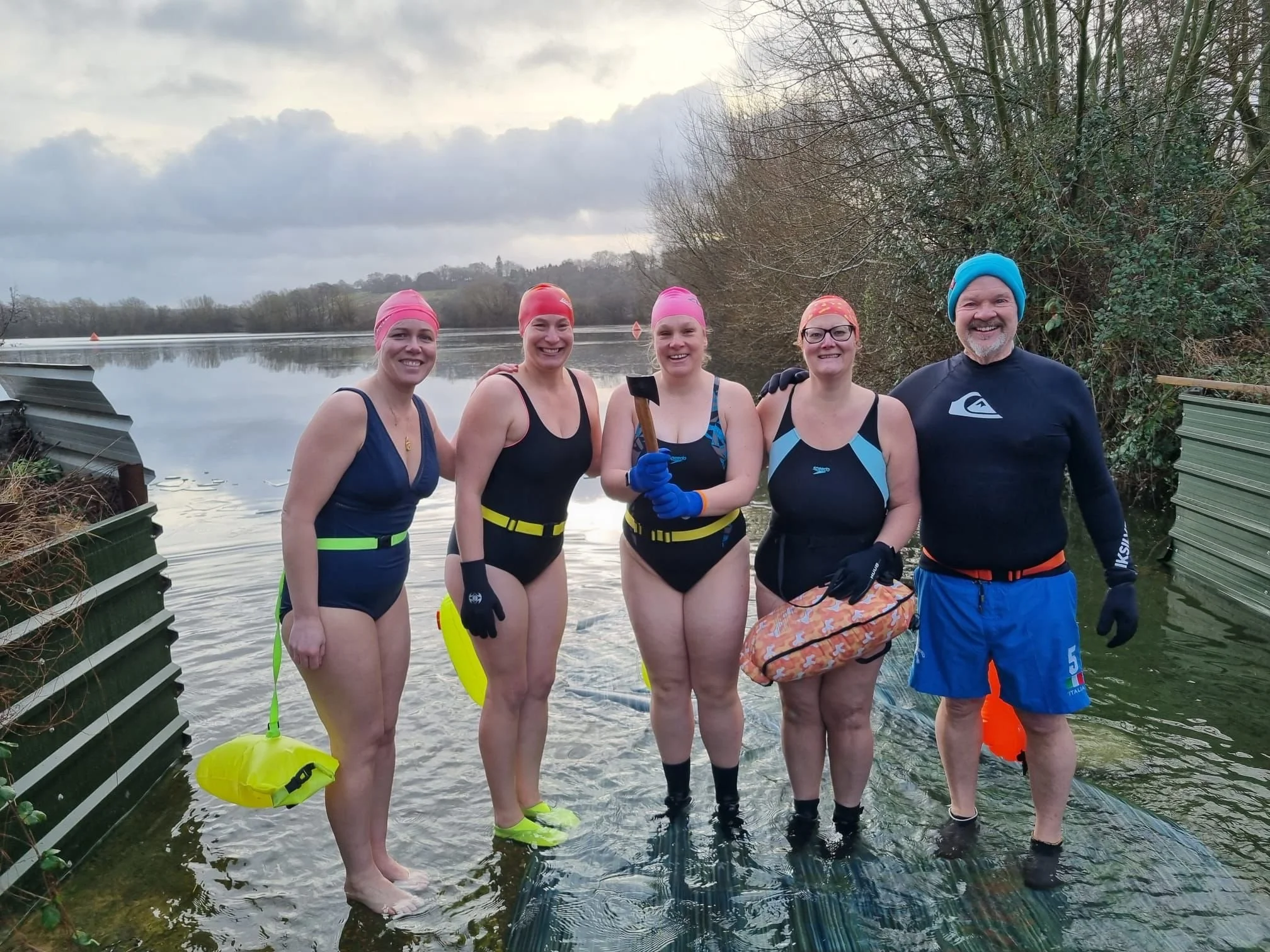 Group of five swimmers standing in a river, wearing swimsuits, pink swim caps, and holding rescue gear. The background features a cloudy sky, leafless trees, and a river with calm water.