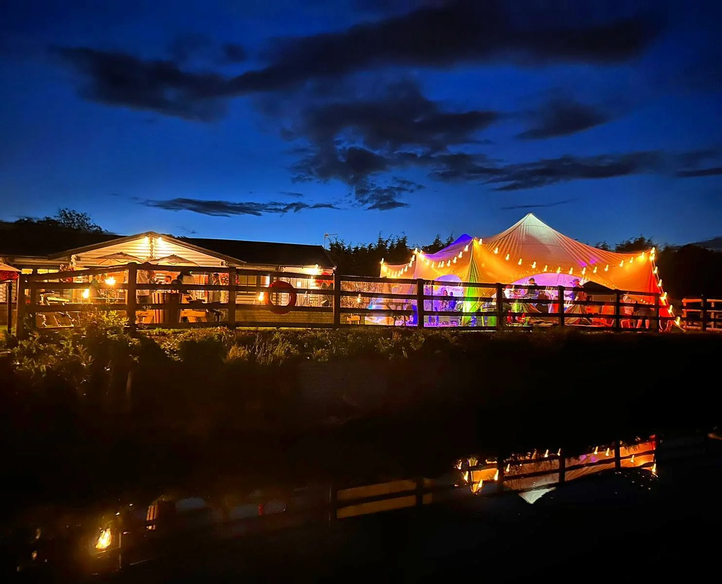 Nighttime outdoor party scene with a large illuminated tent decorated with string lights, a smaller building to the left with people inside, and a cloudy sky above. The setting is reflected in a body of water at the bottom of the image.