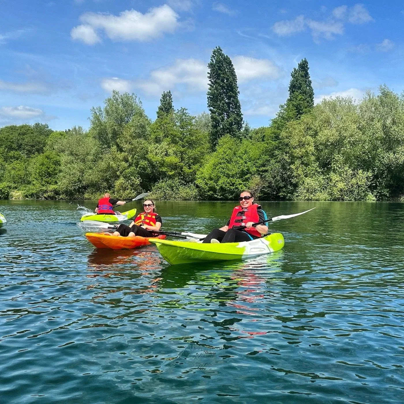 Three women kayaking on a river with green trees and a blue sky in the background.