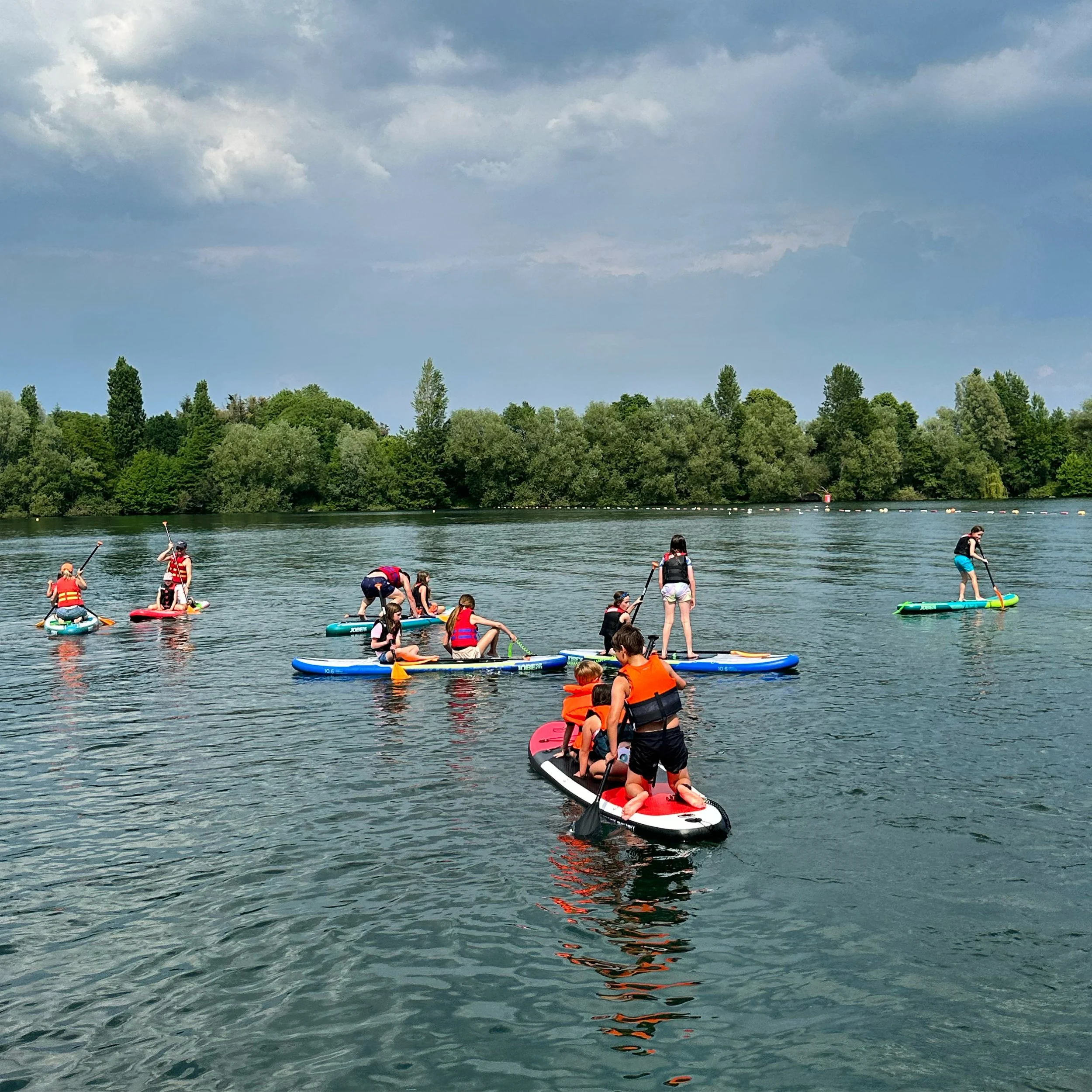 People paddleboarding on a lake on a partly cloudy day, with a forested shoreline in the background.