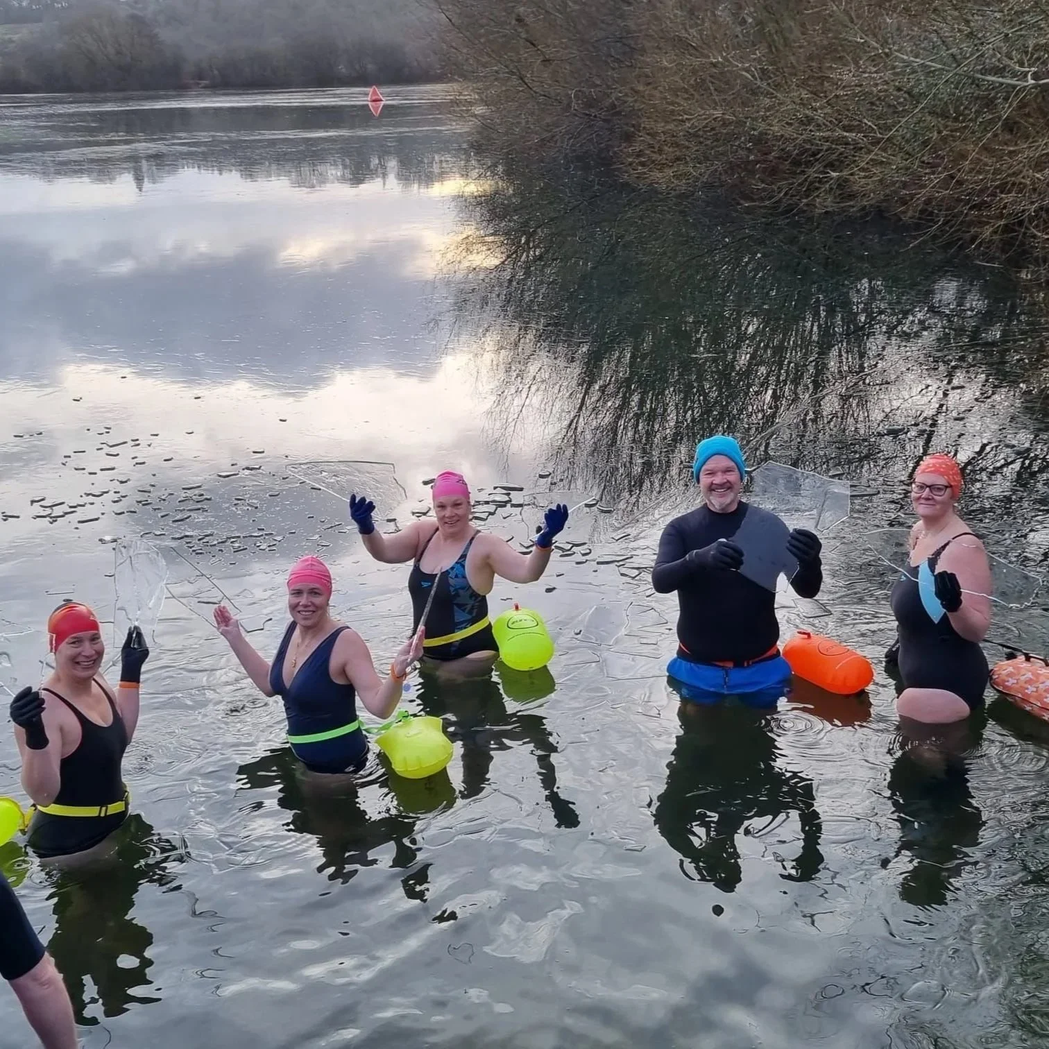 Five people wearing swimsuits, gloves, and colorful swim caps standing in a river, holding broken glass and smiling.