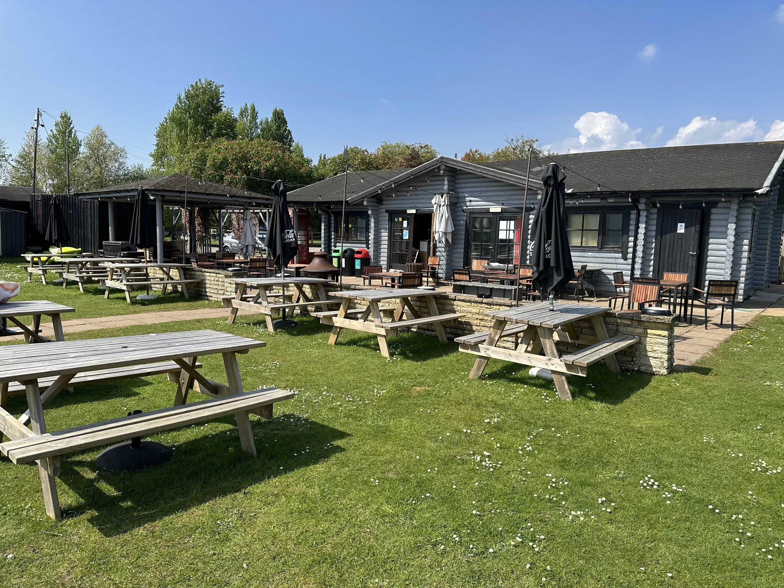 Outdoor patio area with wooden picnic tables, black umbrellas, and a building with gray log-style siding under a blue sky with some clouds.
