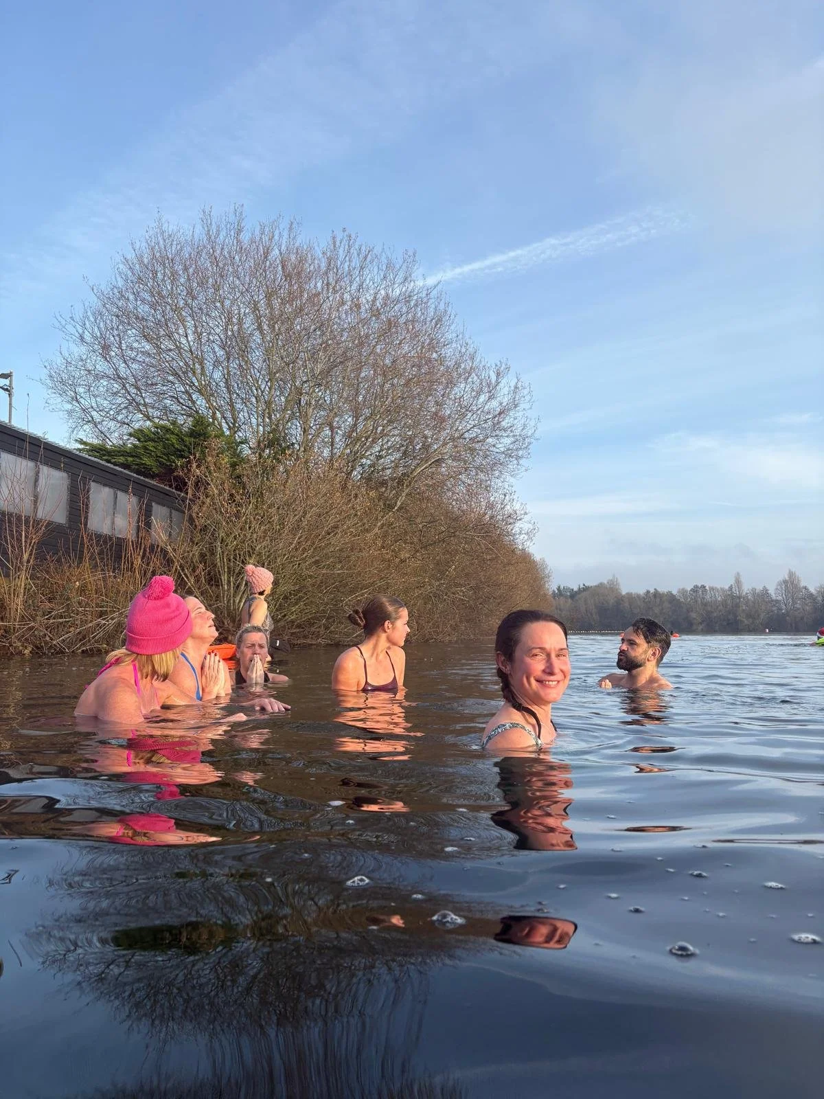Group of people swimming and relaxing in a natural body of water on a cloudy day with trees in the background.