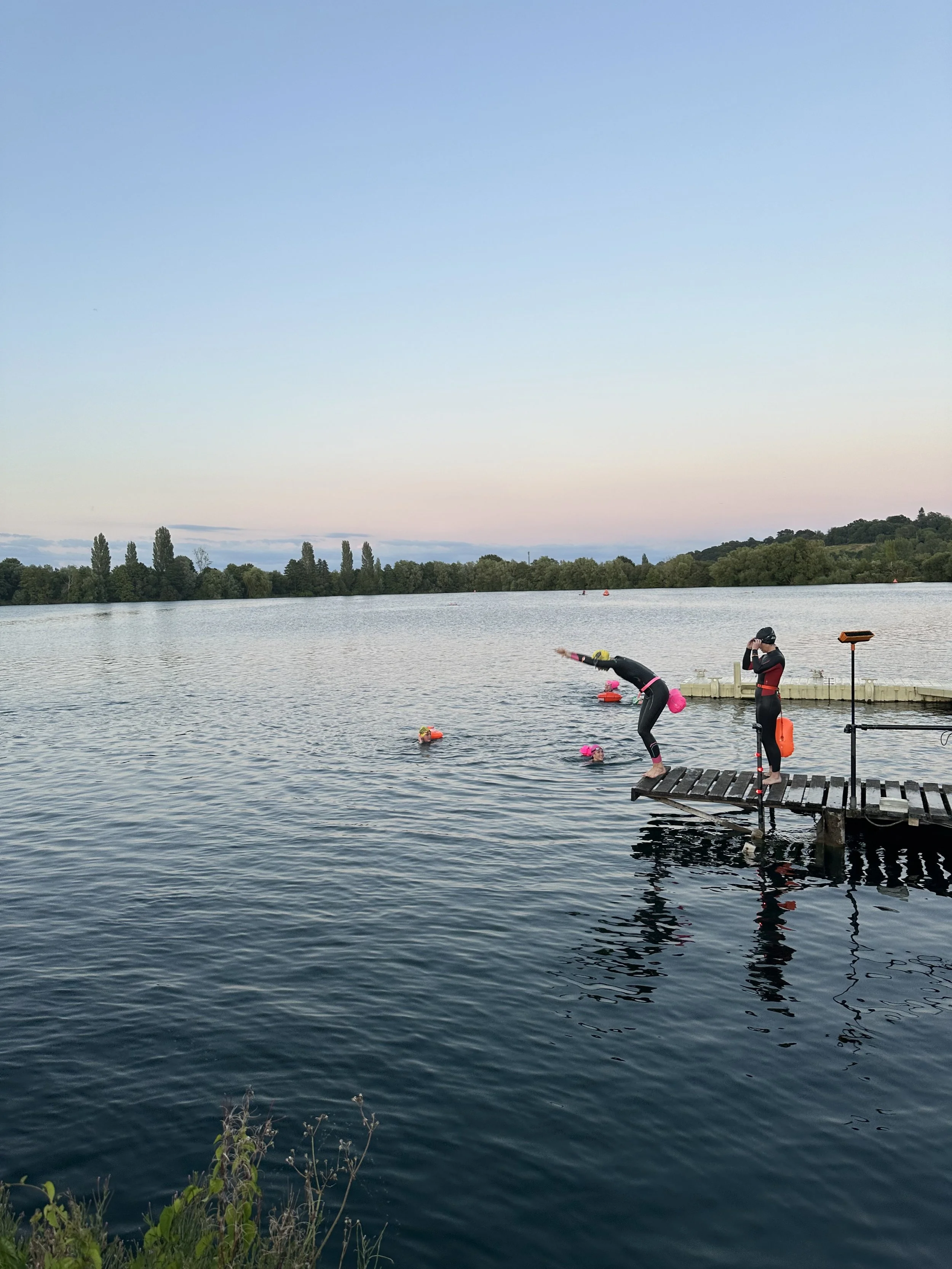 People in wetsuits preparing to swim from a dock into a lake during dawn or dusk.