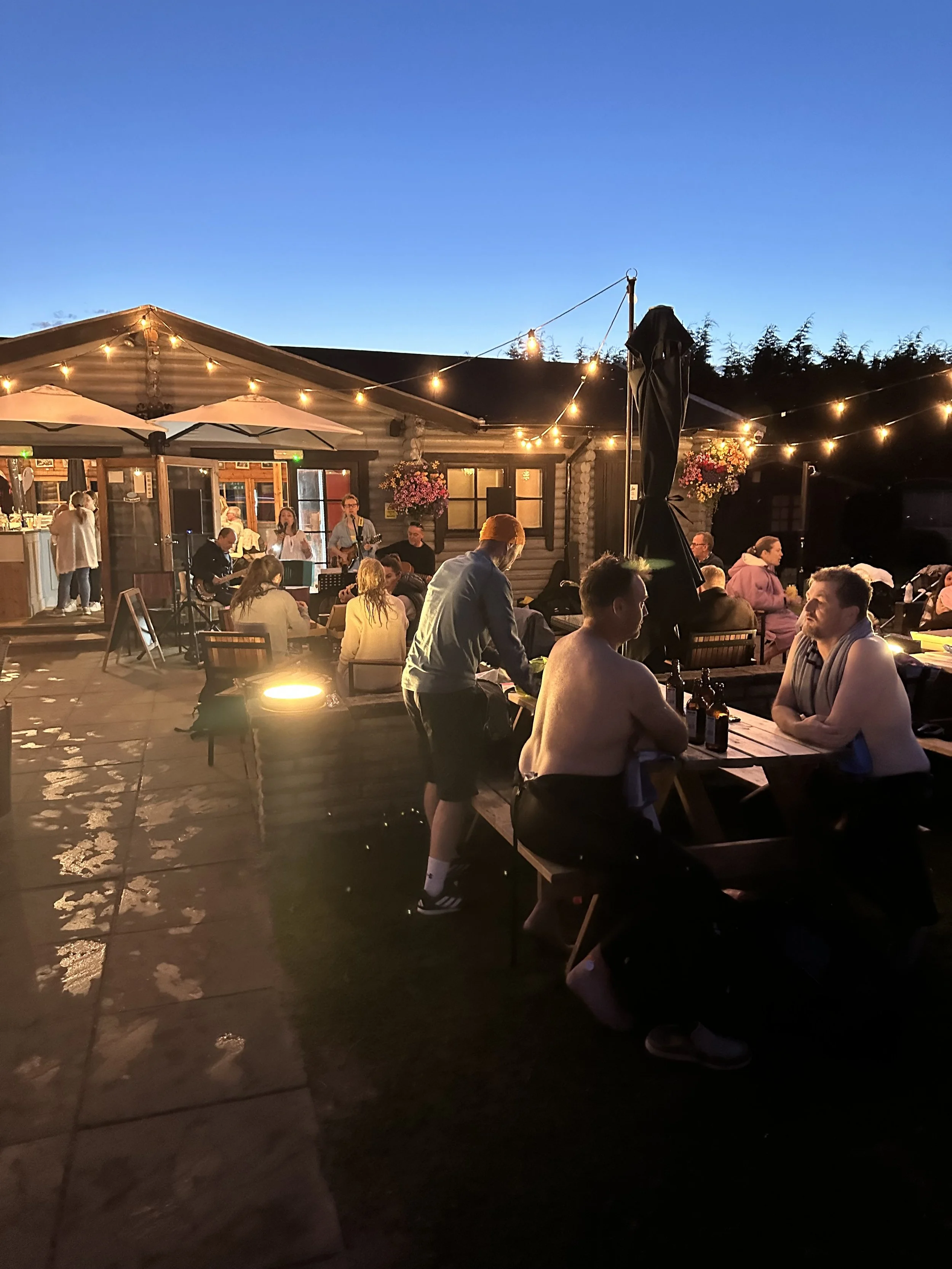 Outdoor evening gathering with people sitting and chatting at picnic tables, some shirtless, under string lights near a rustic building with a band playing in the background.