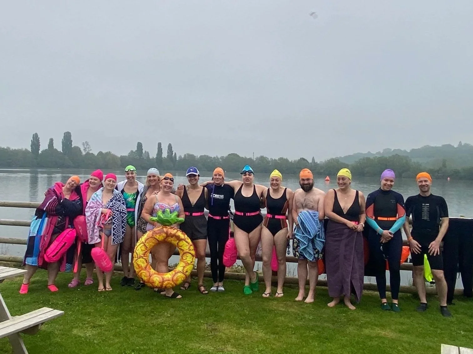 A group of people in swimsuits and swim caps standing together outdoors near a body of water, smiling for a photo.