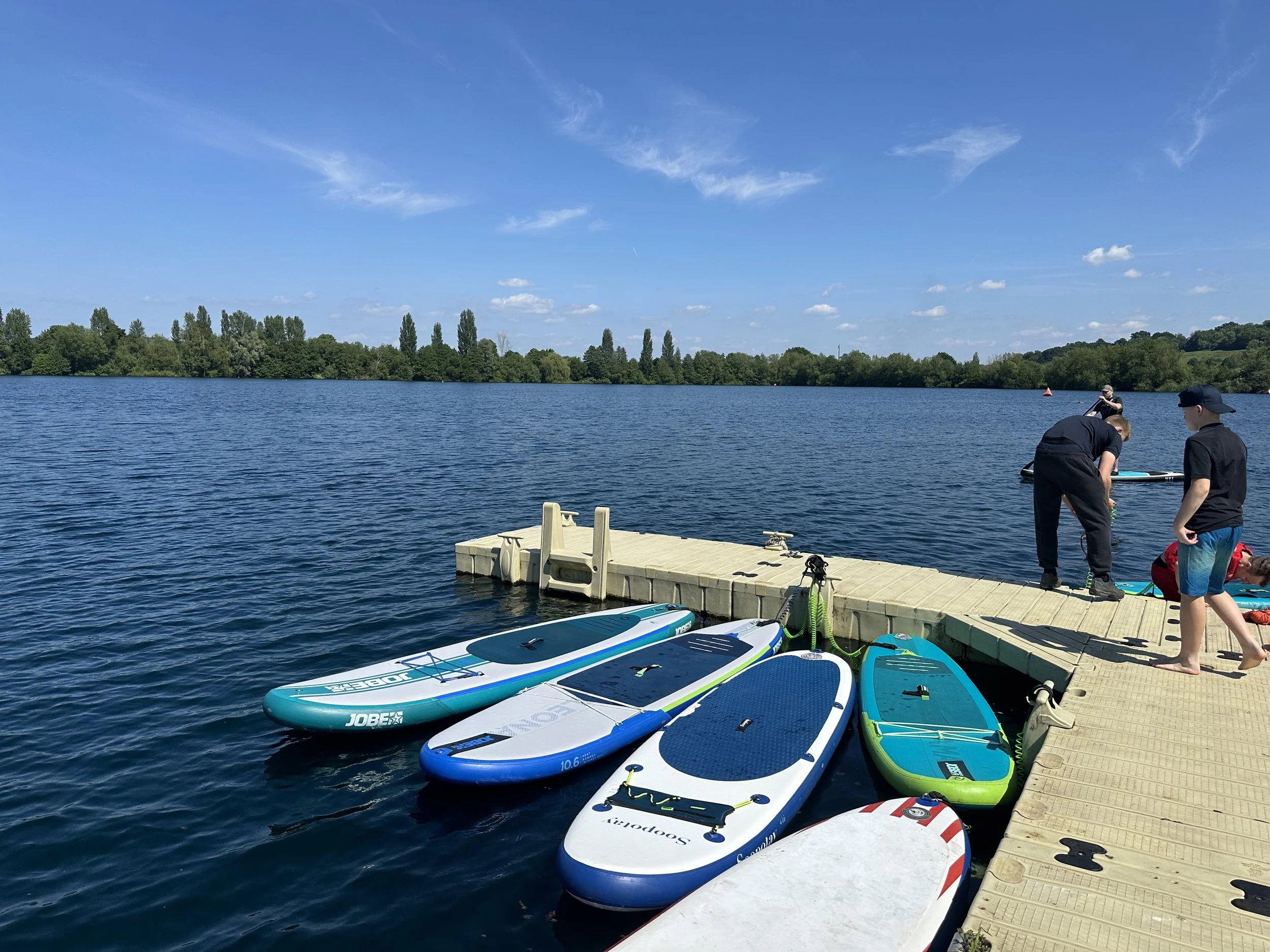 People preparing paddleboards and kayaks on a dock by a large lake on a sunny day with blue skies and greenery in the background.