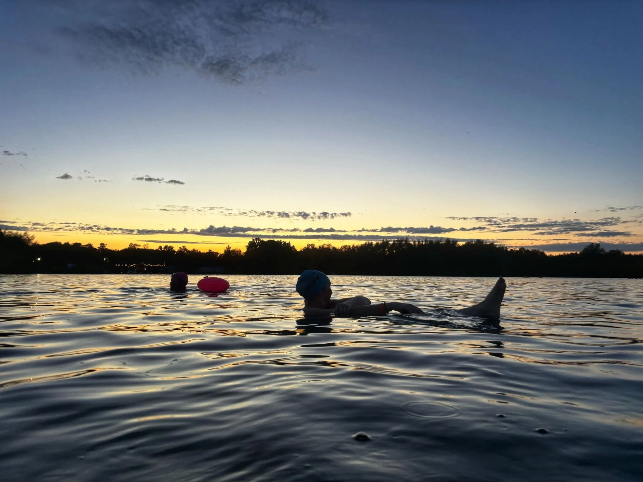 Person swimming in a lake during sunset, wearing a blue swim cap, with two other swimmers visible in the background, and trees along the shoreline.