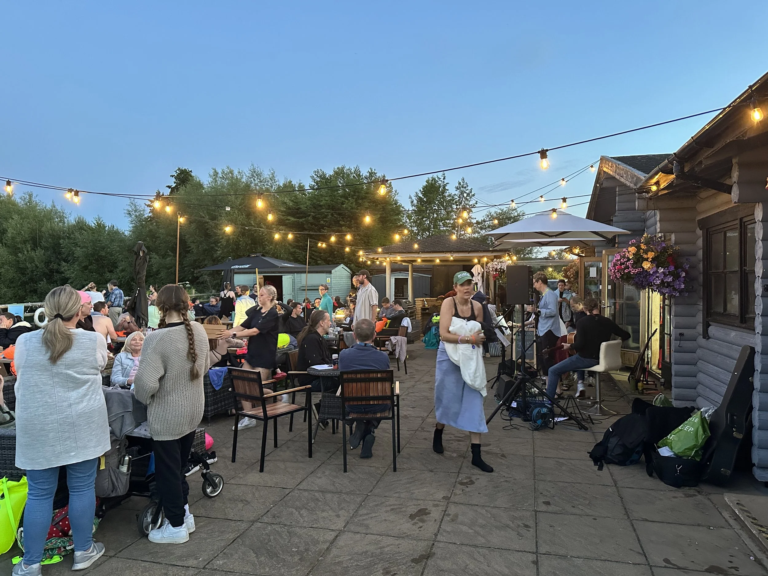 Outdoor gathering with people socializing and enjoying live music, illuminated by string lights, on a patio with logs and flowers.