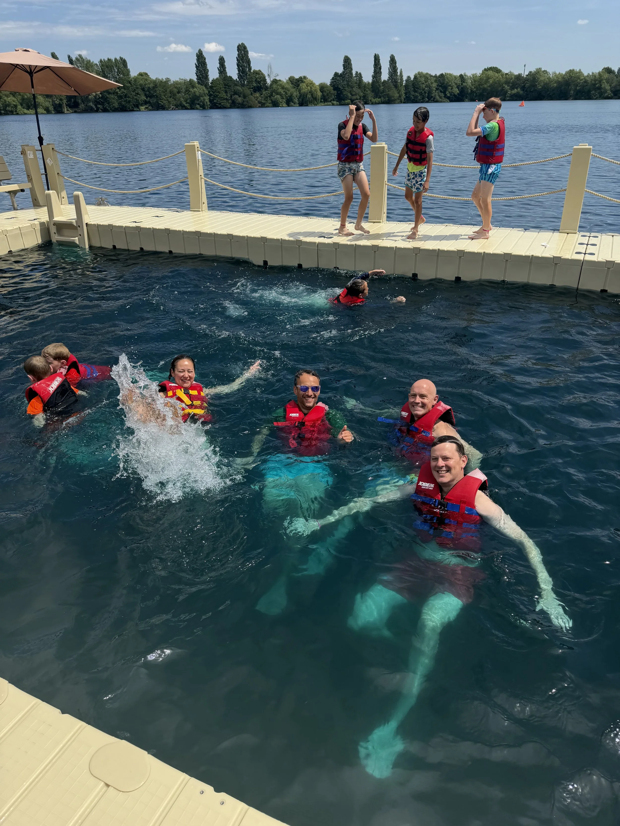 Children and adults wearing life jackets swimming and playing in a lake near a floating dock with a rope barrier, under a partly cloudy sky.