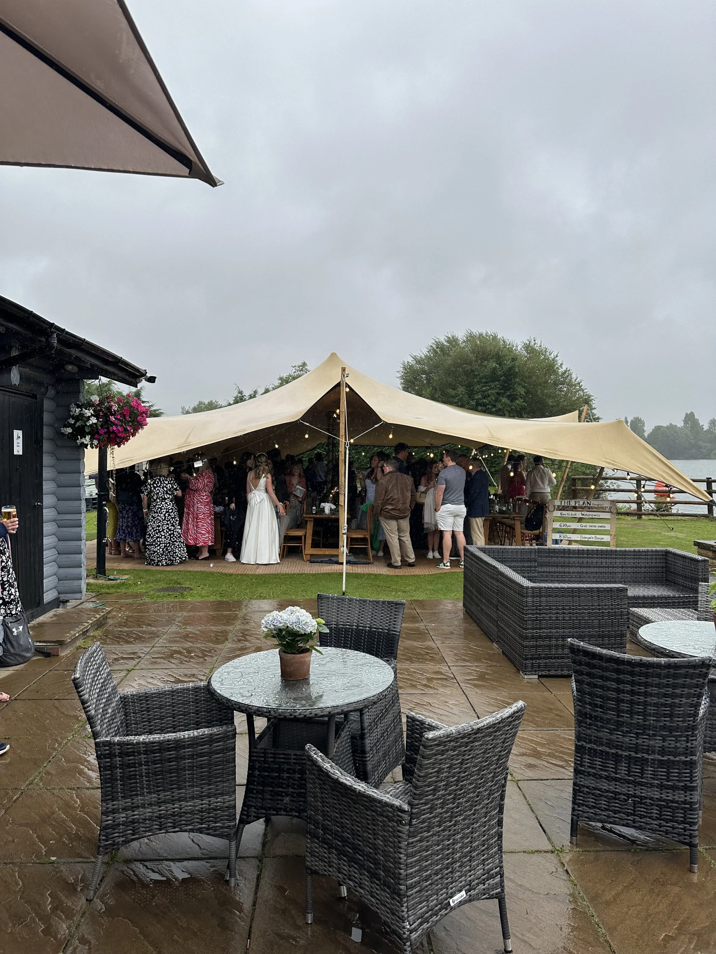 Outdoor gathering under a beige tent with people socializing, on a rainy day with wet patio and cloudy sky, near a lake with trees in background.