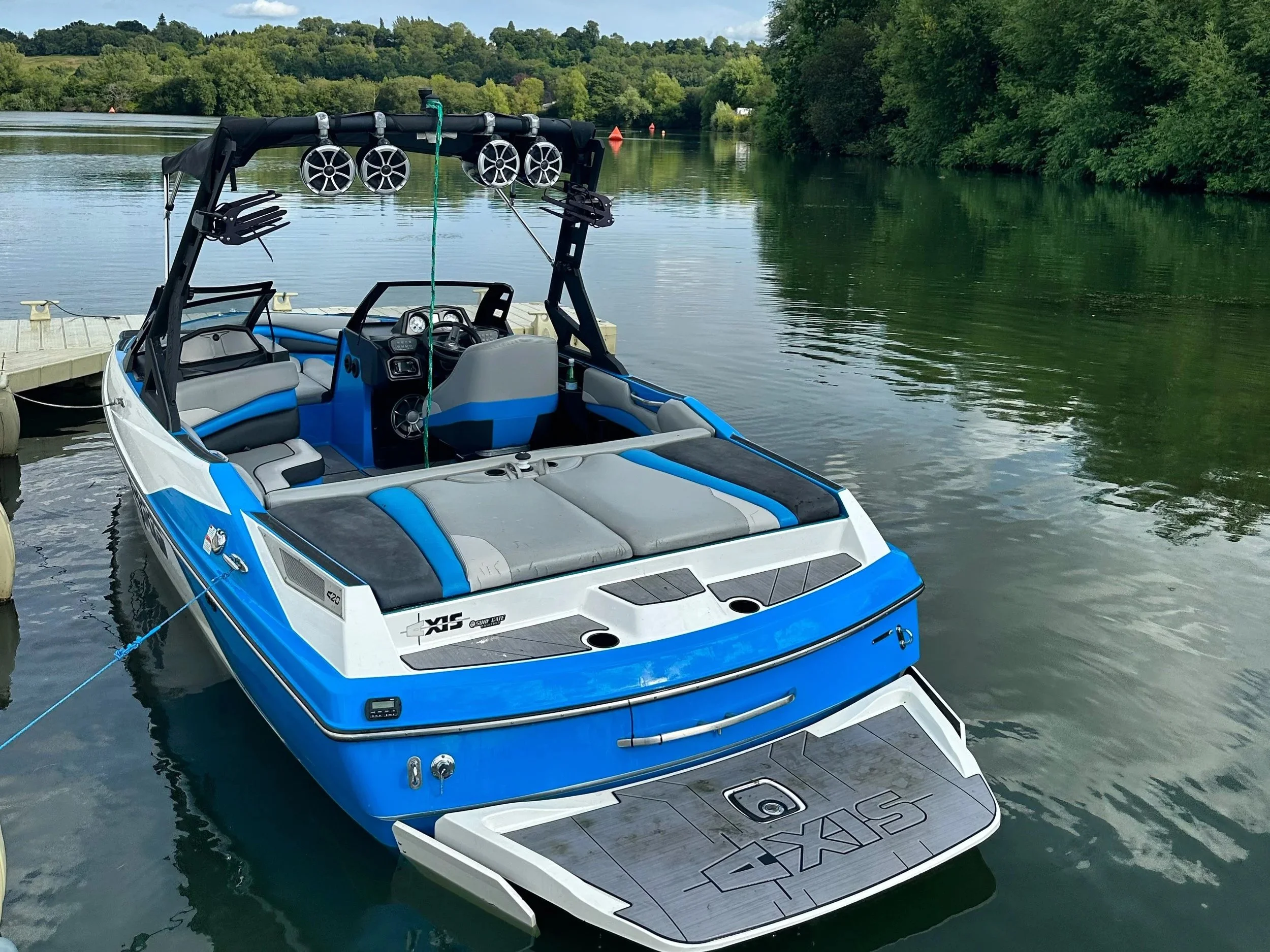 A blue and white luxury speedboat docked on a calm lake surrounded by green trees. The boat features a modern design with seating, a steering wheel, and an extended swim platform at the back.