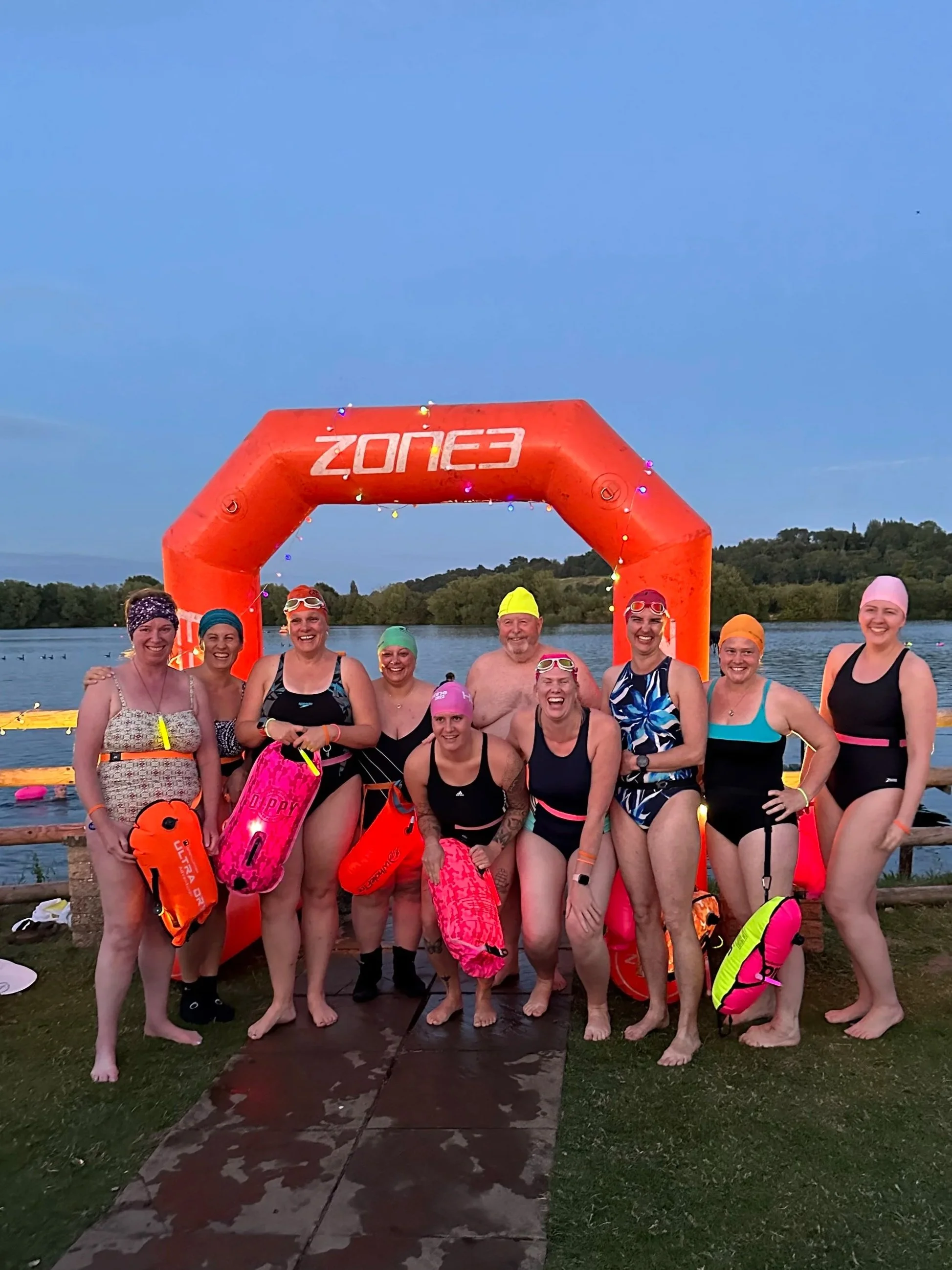 Group of people in swimsuits and swim caps standing at the poolside with lake in the background, under an orange inflatable arch labeled 'ZONE3' during sunset or early evening.