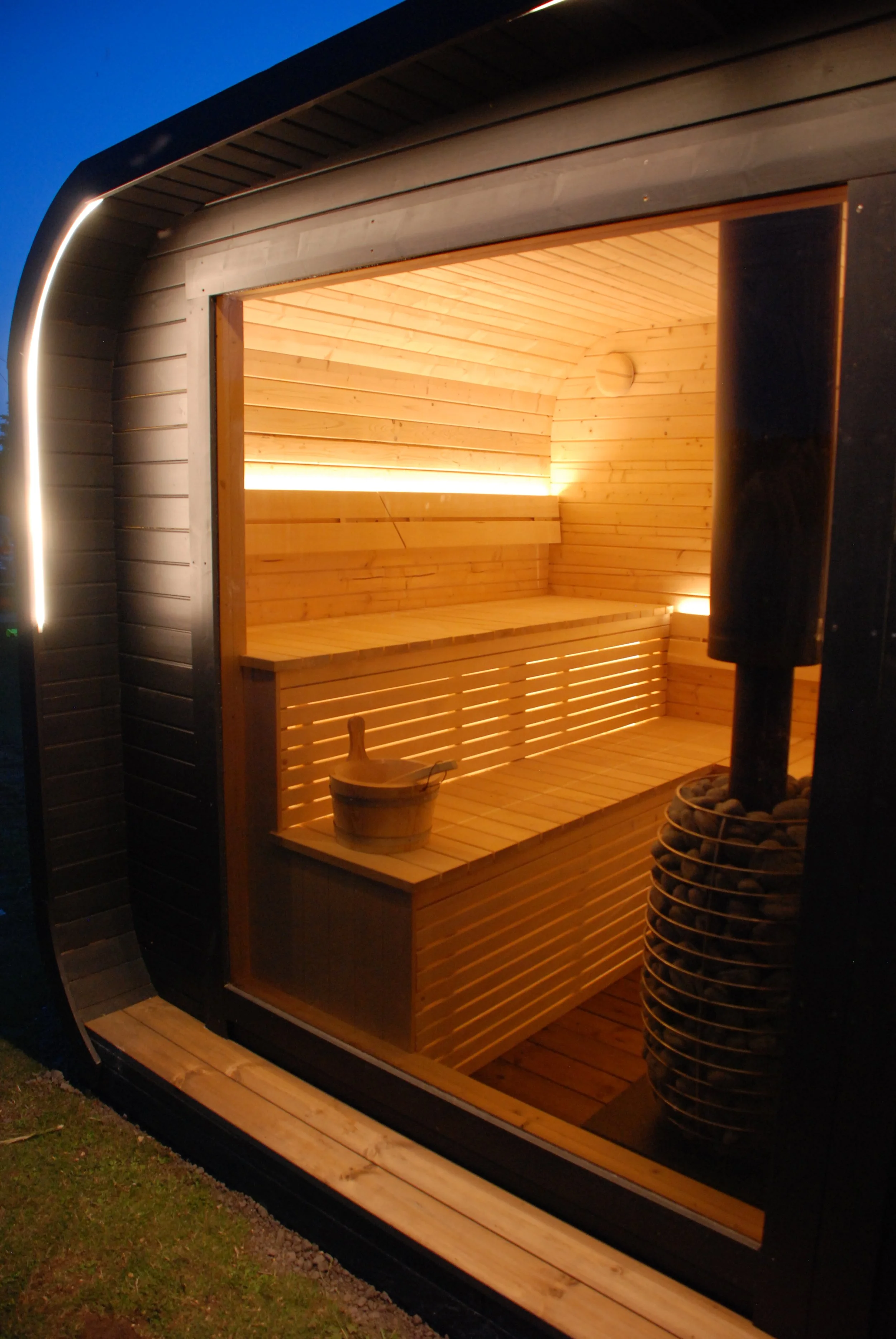 Inside a modern outdoor sauna with wooden benches and interior lighting, viewed through a glass door.
