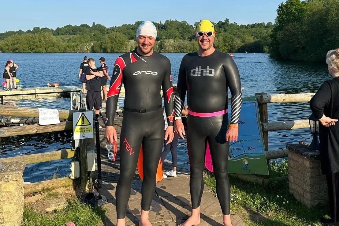 Two men in wetsuits standing on a dock by a lake, smiling at the camera, with other swimmers in the background preparing for a swim event.