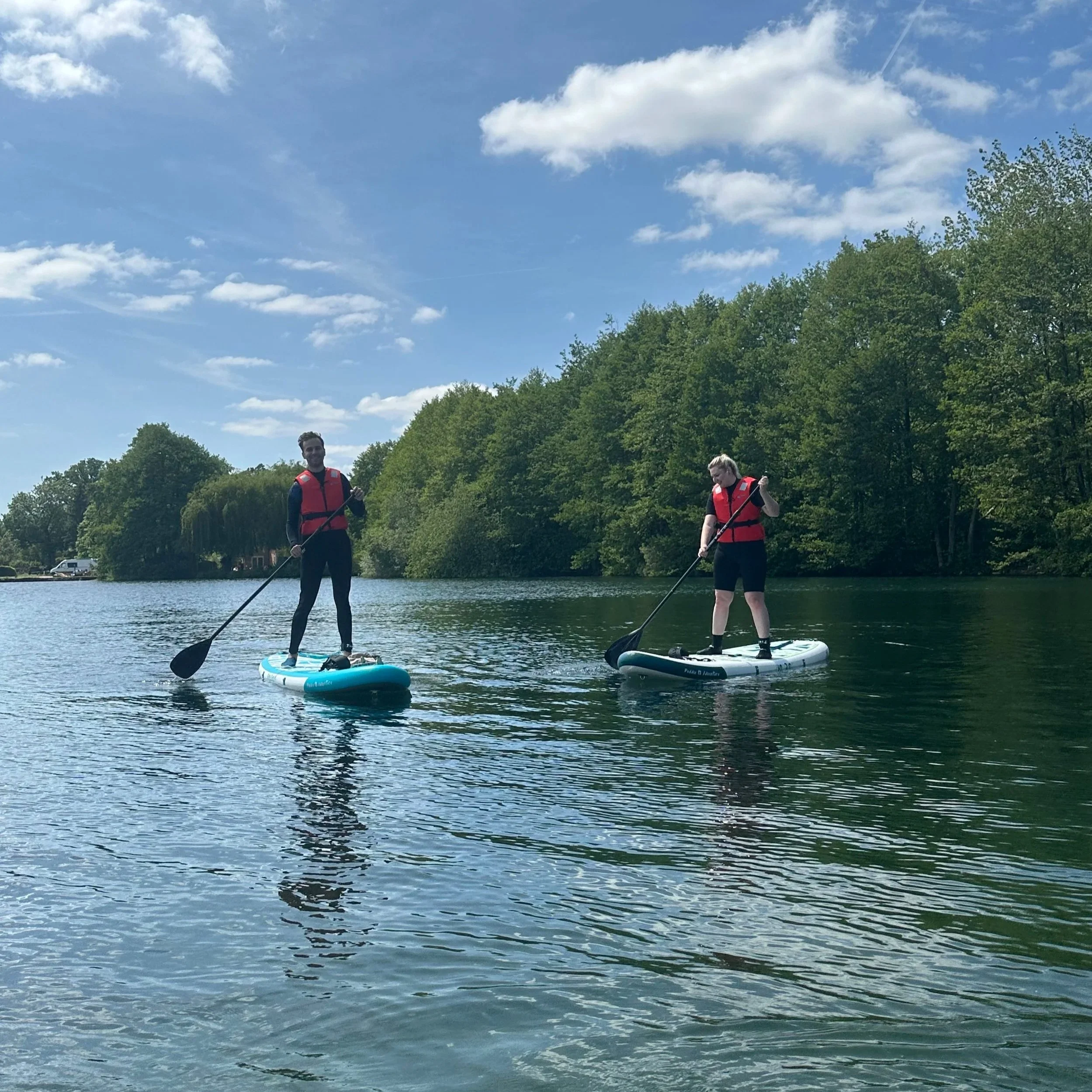 Two people paddleboarding on a lake under a blue sky with scattered clouds, surrounded by green trees.