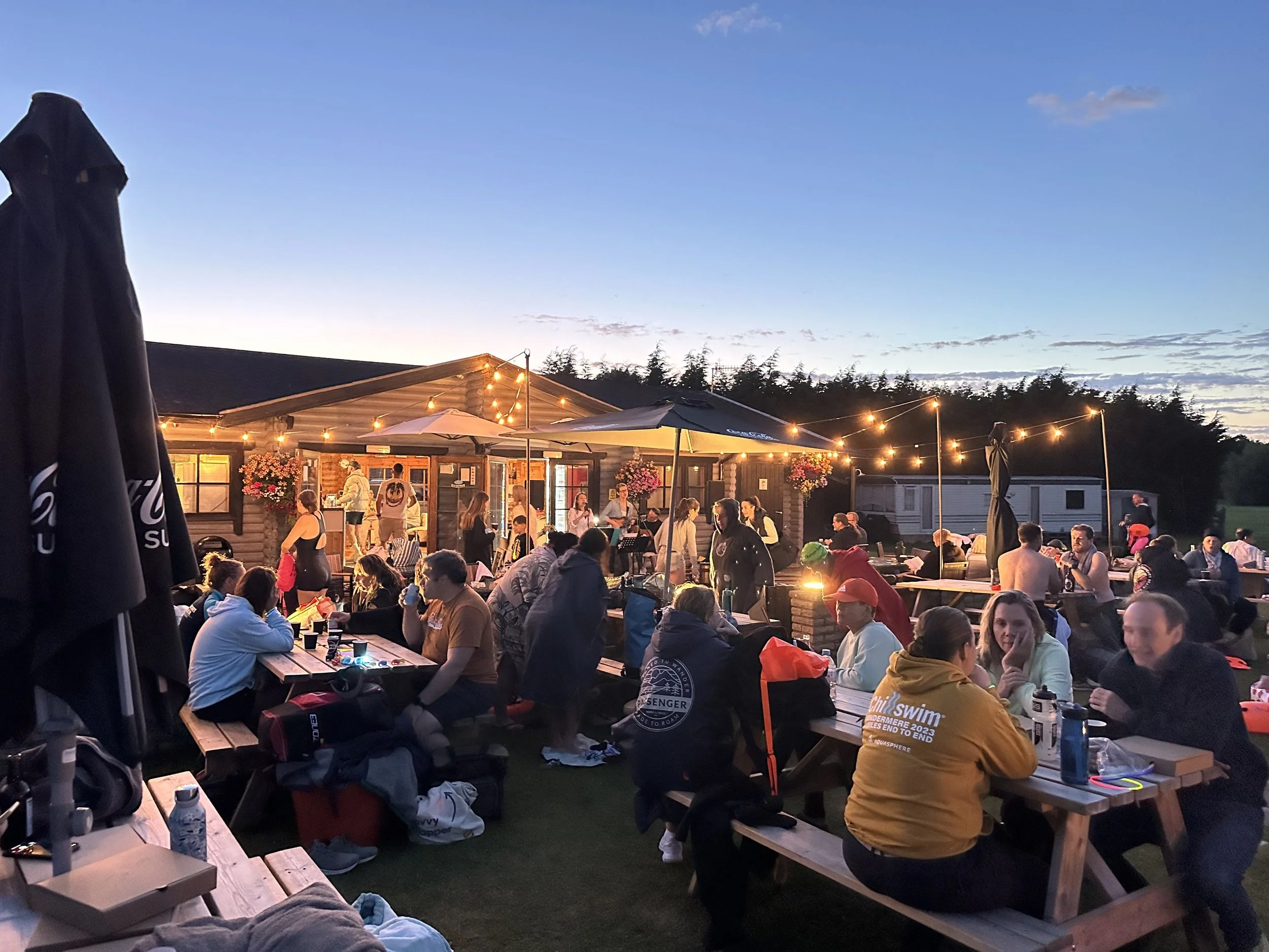 People gathered at an outdoor evening event at a picnic area with tables, string lights, umbrellas, and a building in the background, enjoying food and drinks.