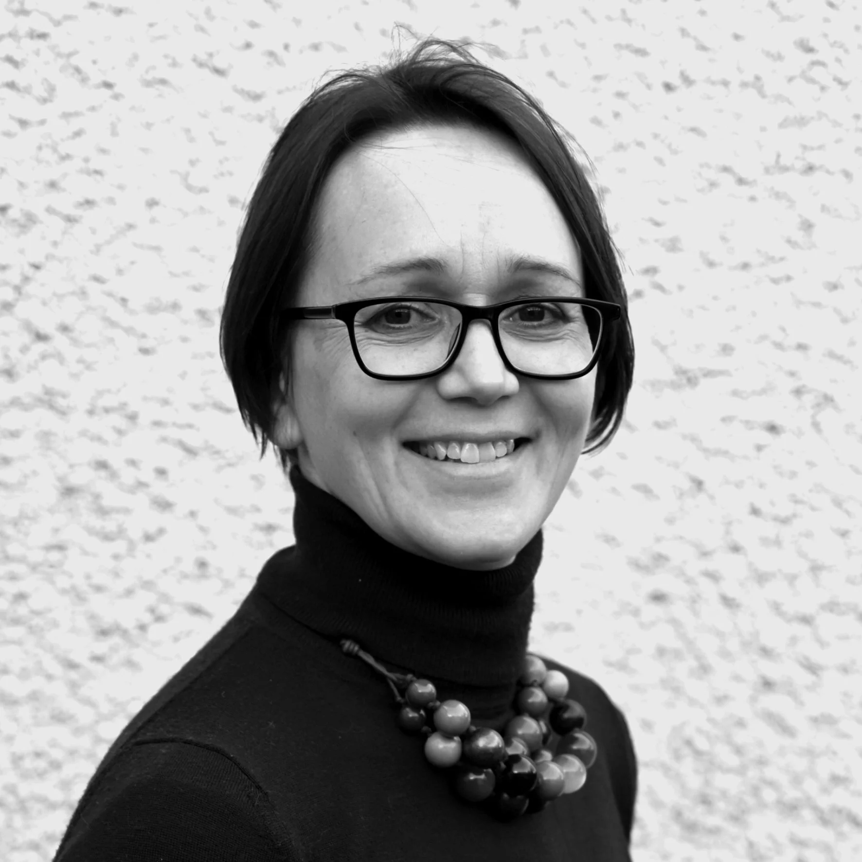 Black and white photo of a Mel Dixon with short hair, wearing glasses, a turtleneck, and a necklace, smiling at the camera against a textured background.