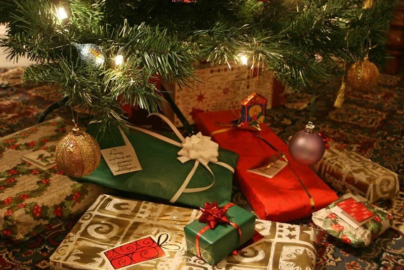Under a decorated Christmas tree are wrapped gifts, including a gold, green, red, and patterned presents with ribbons and bows, on a festive carpet.