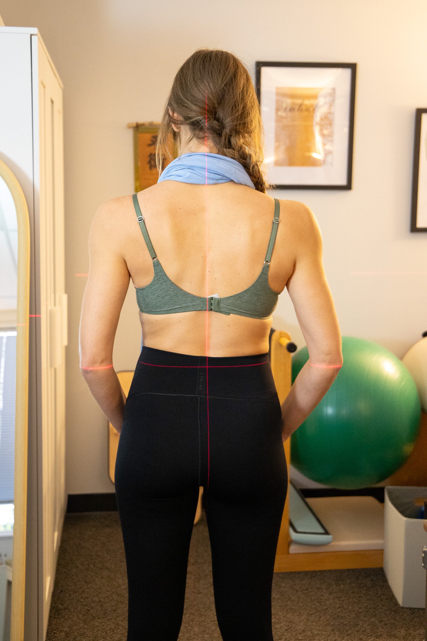 Back view of a woman with long brown hair in a braid, wearing a gray sports bra, black high-waisted pants, and a light blue scarf around her neck, standing in a room with exercise equipment including an exercise ball and a mirror.