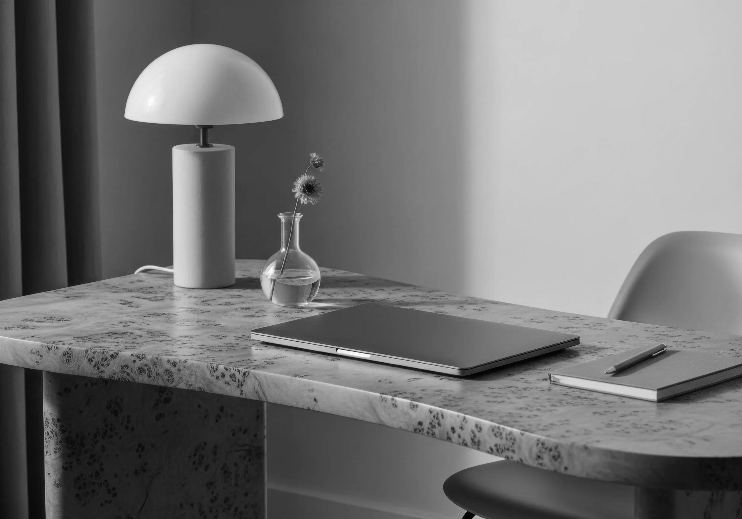 Black and white photo of a marble table with a modern lamp, a small glass vase with flowers, a closed laptop, a notebook, and a pen.