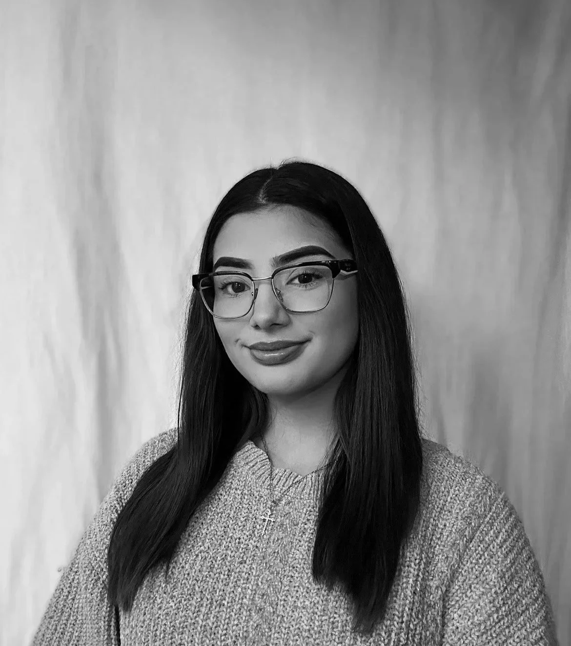 A young woman with long dark hair and glasses, smiling and wearing a knit sweater, a necklace, and lipstick, standing in front of a plain background.