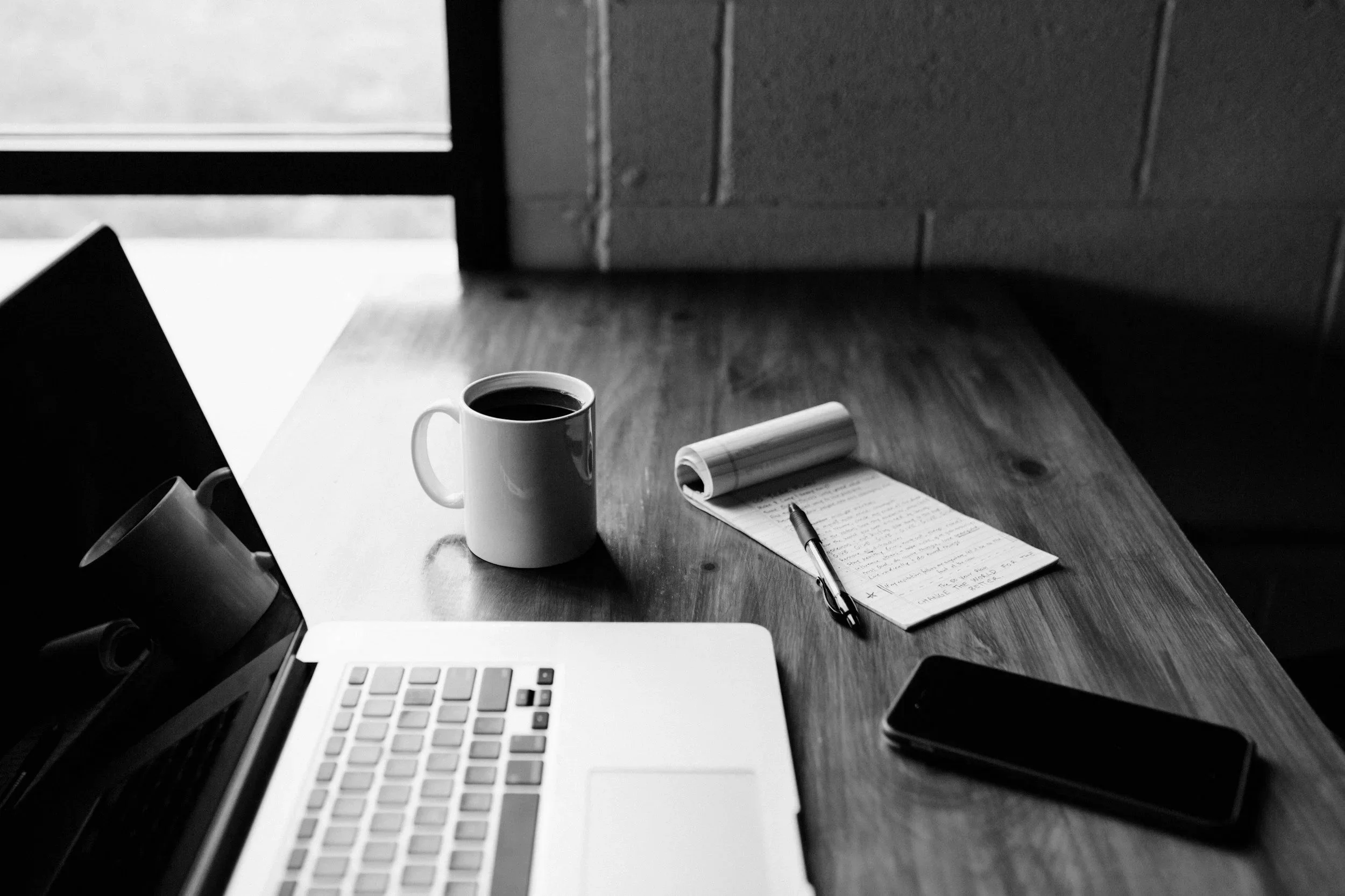 Black and white photo of a workspace on a wooden table with a laptop, a coffee mug, a pen, a notebook, and a smartphone.