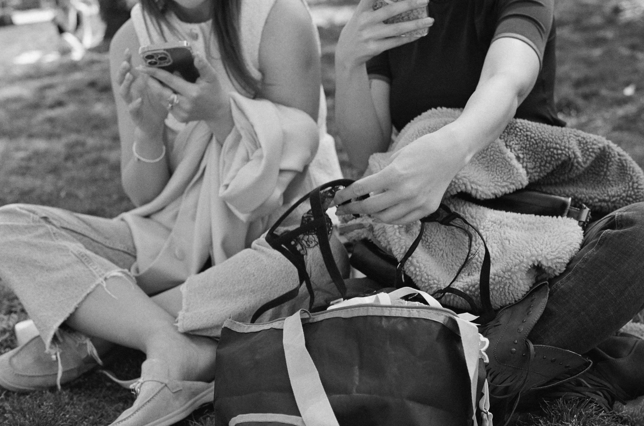 Two women sitting on the grass outdoors; one is holding a phone, the other is reaching for a backpack.
