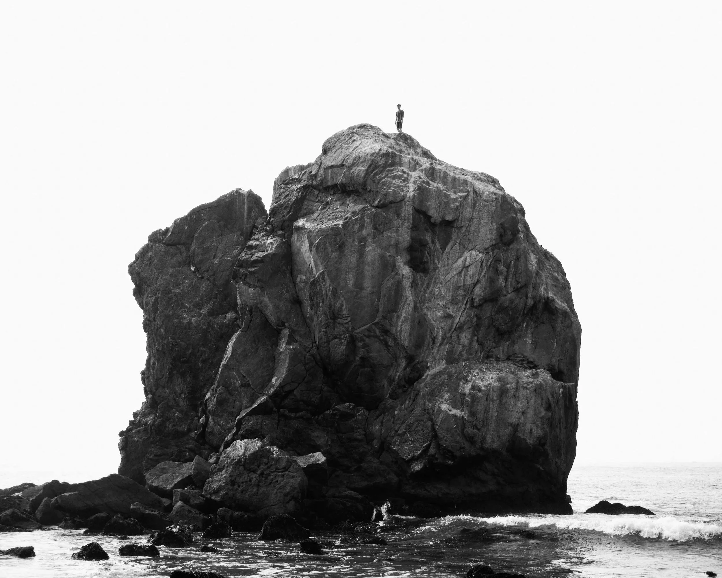 A person standing atop a large rocky formation in the ocean, black and white photo.