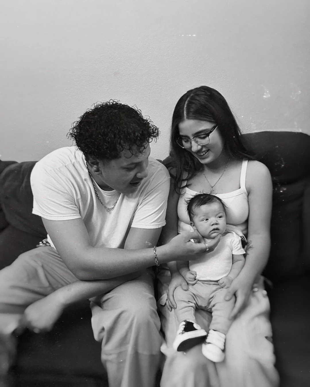 A black and white photo of a man and woman sitting on a couch with a baby, smiling and looking at the baby, in a cozy indoor setting.