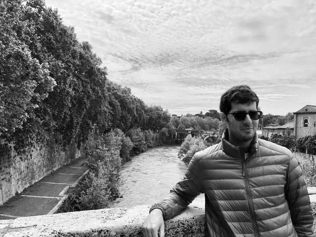 A young man wearing sunglasses and a puffer jacket standing on a bridge over a river, with trees on the riverbank and buildings in the background, in a grayscale outdoor scene.