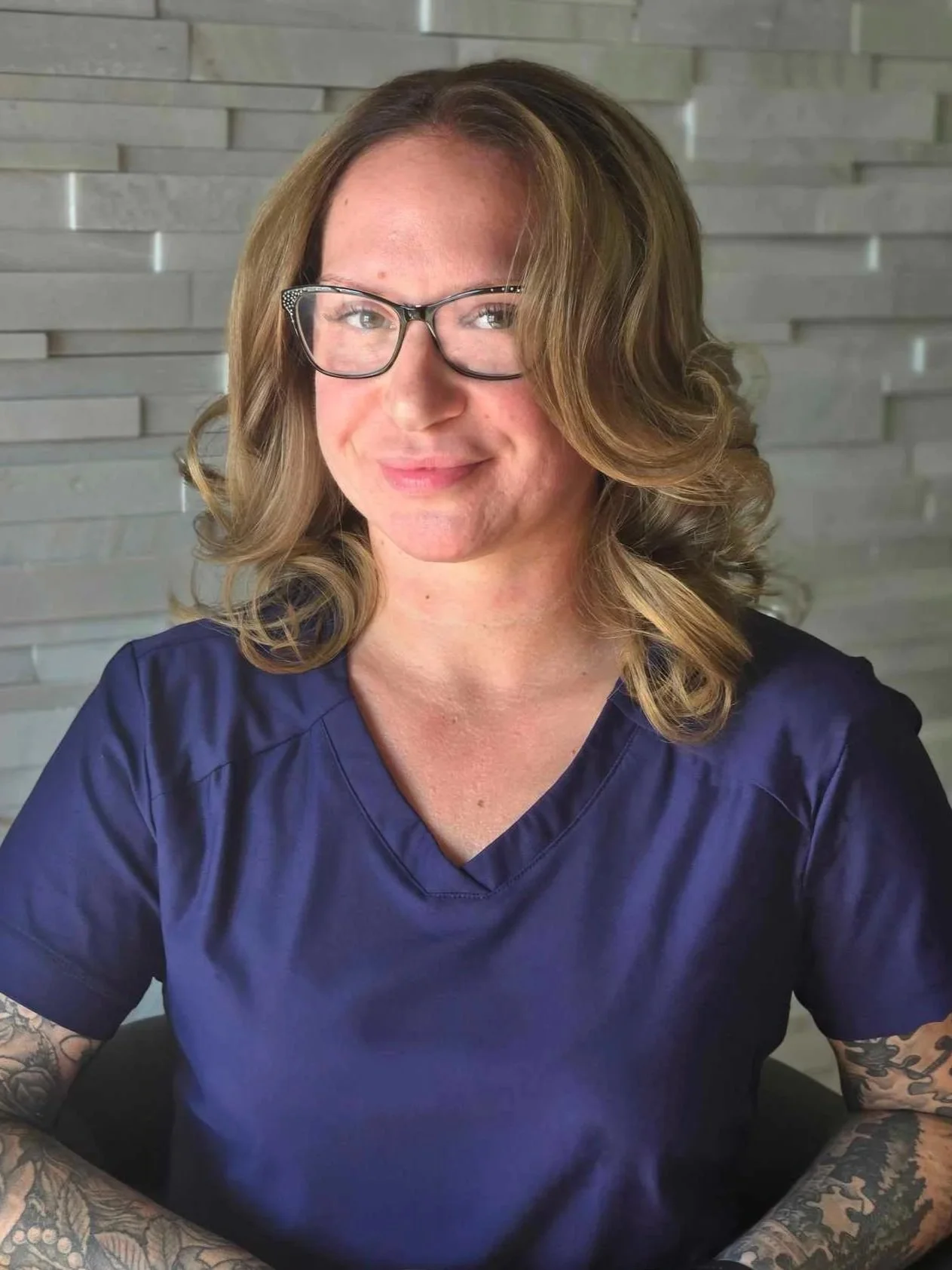 A woman with shoulder-length wavy blonde hair, wearing glasses and a navy blue top, smiling at the camera in front of a gray textured wall.