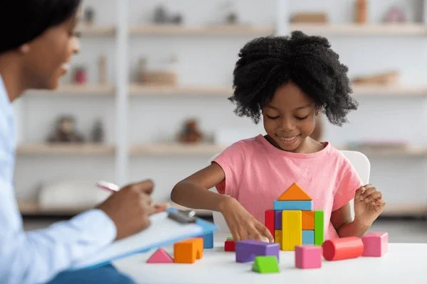 A young girl playing with colorful building blocks at a table, with an adult woman sitting nearby taking notes.