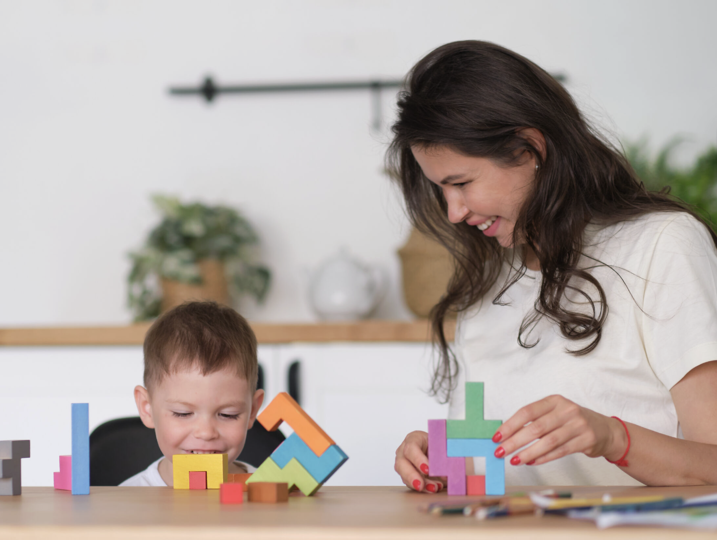 A woman and a young boy playing with colorful geometric wooden blocks at a table in a bright, modern room.