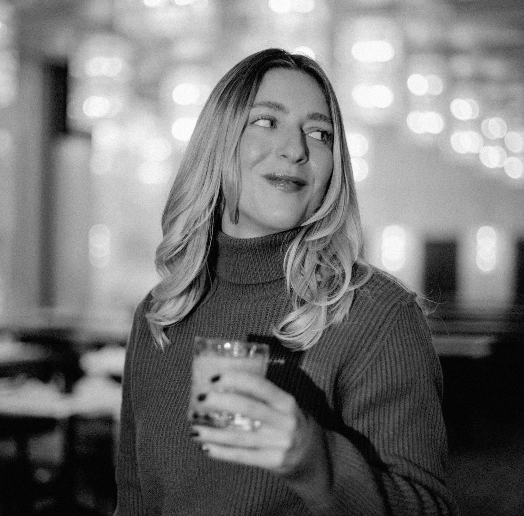 Frau mit lockigem Haar trägt Rollkragenpullover, hält ein Glas, und lächelt, im Hintergrund geschäftliche Beleuchtung.