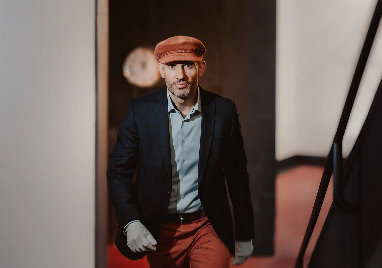 Ein Mann in formeller Kleidung mit schwarze Jacke, blauer Hemd, braune Hose, trägt einen braunen Hut und einen weißen Handschuh, steht in einem Raum mit dunklem Hintergrund.