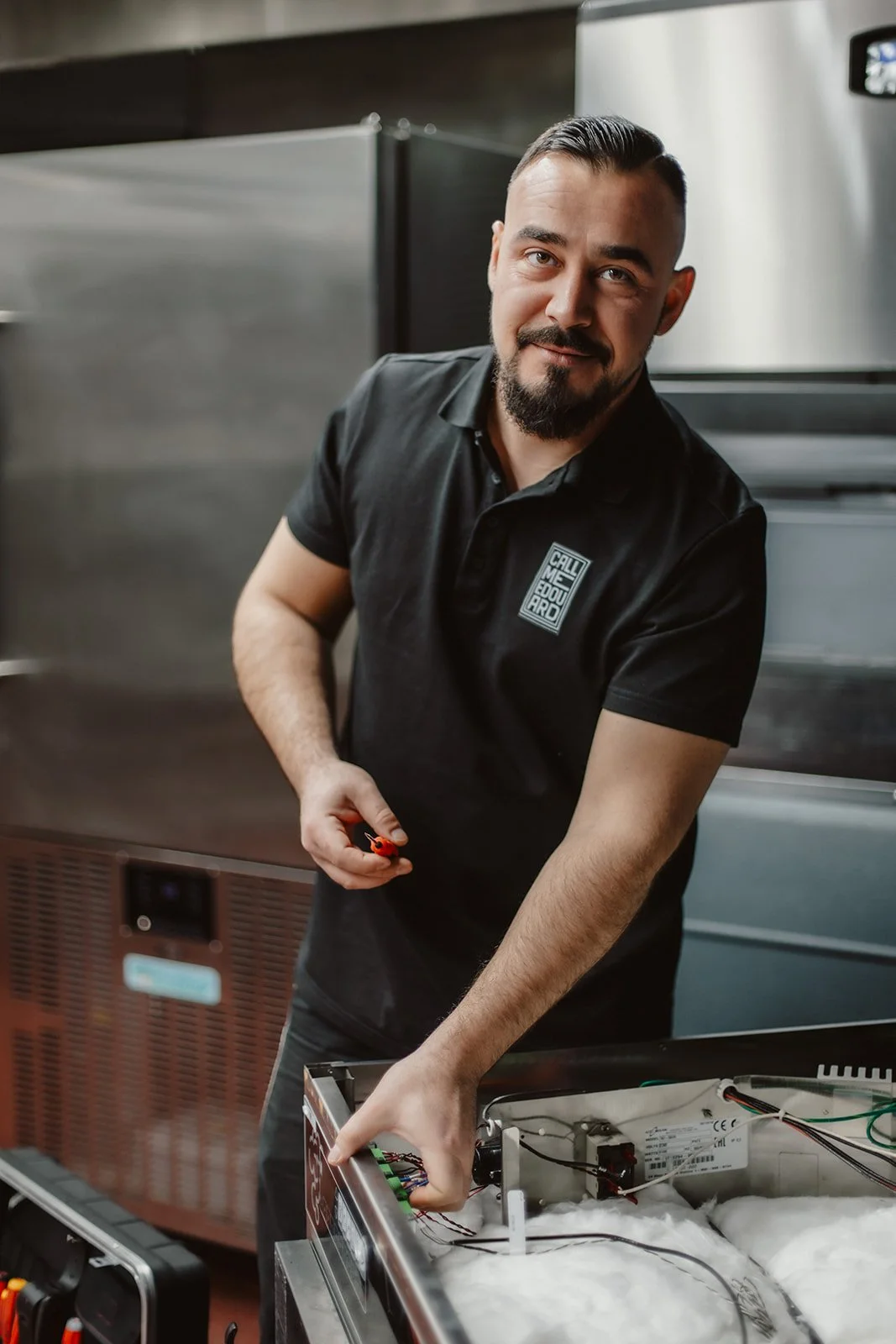 Ein Mann arbeitet an einem technischen Gerät, wahrscheinlich in einer Küche, trägt ein schwarzes Polo-Shirt mit einem Logo und hält ein Werkzeug in der Hand.