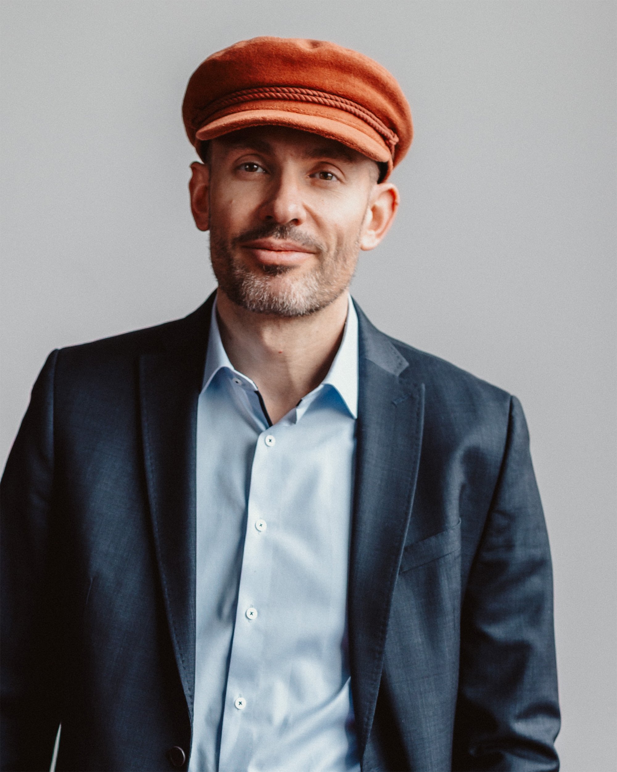 Ein Mann mit grauem Bart, im dunklen Anzug und einer hellblauen Hemd, trägt eine orangefarbene Mütze und steht vor einem grauen Hintergrund.